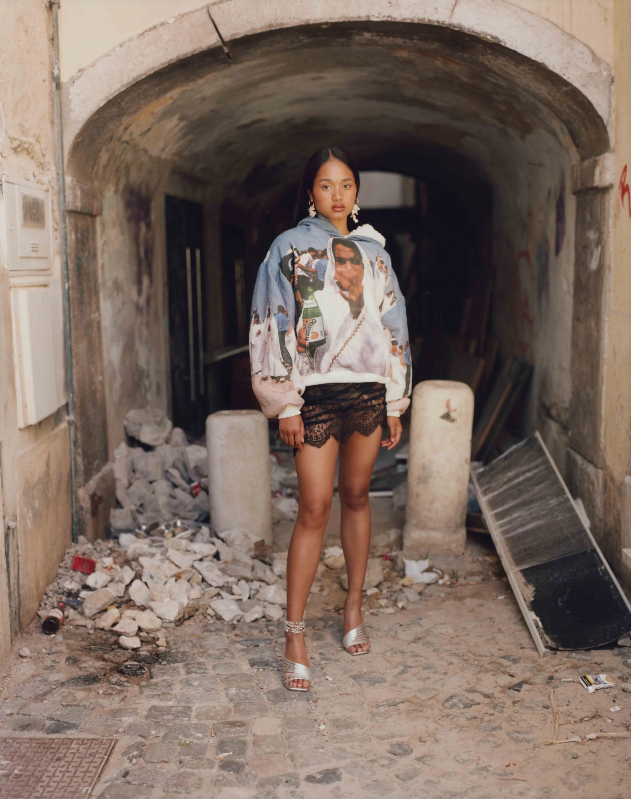 Young woman standing under an arched stone passageway with rubble and debris on the ground, wearing a graphic hoodie, lace shorts, and high heels.