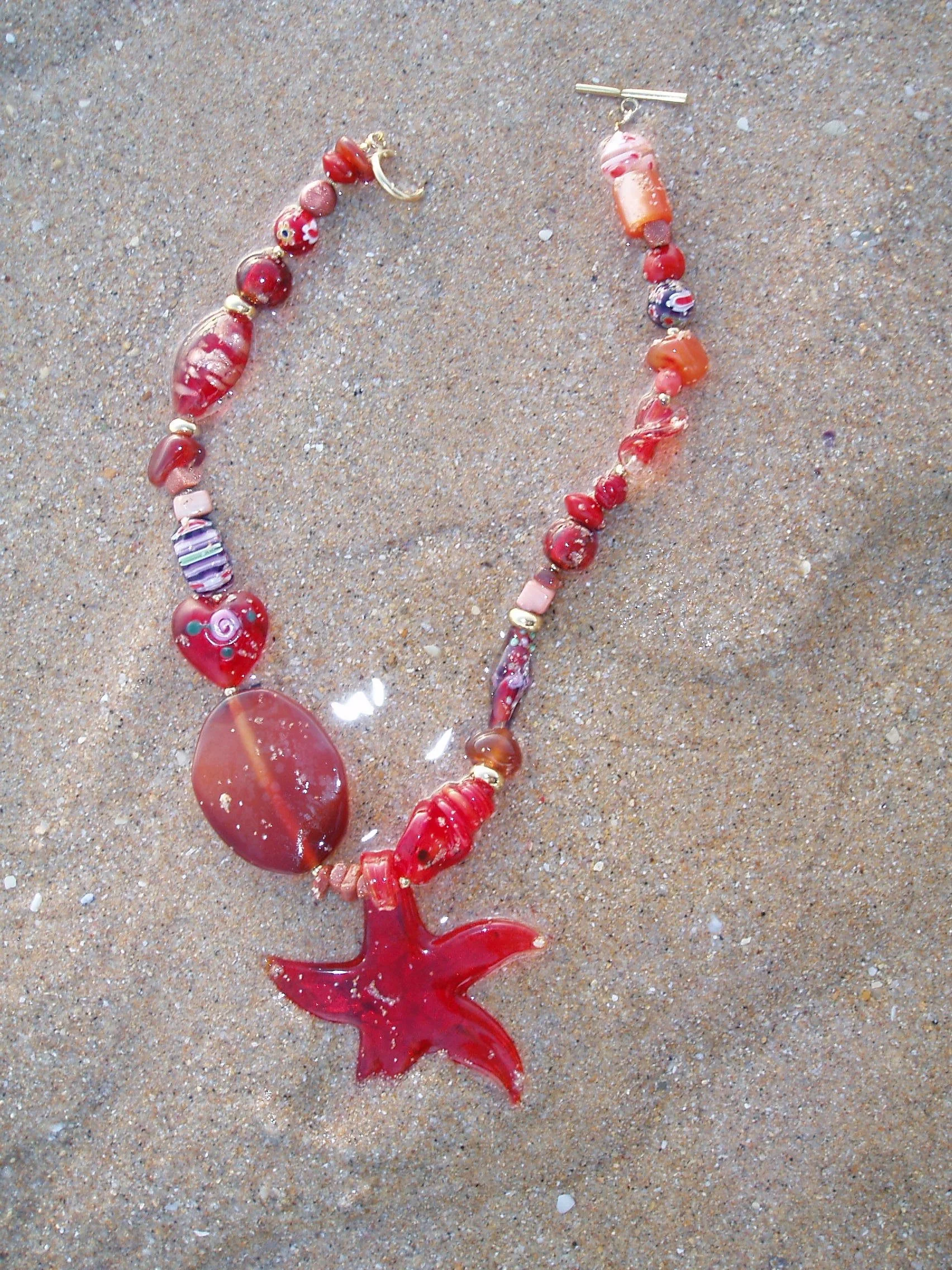 Beaded necklace with a red starfish pendant, lying on sandy surface.