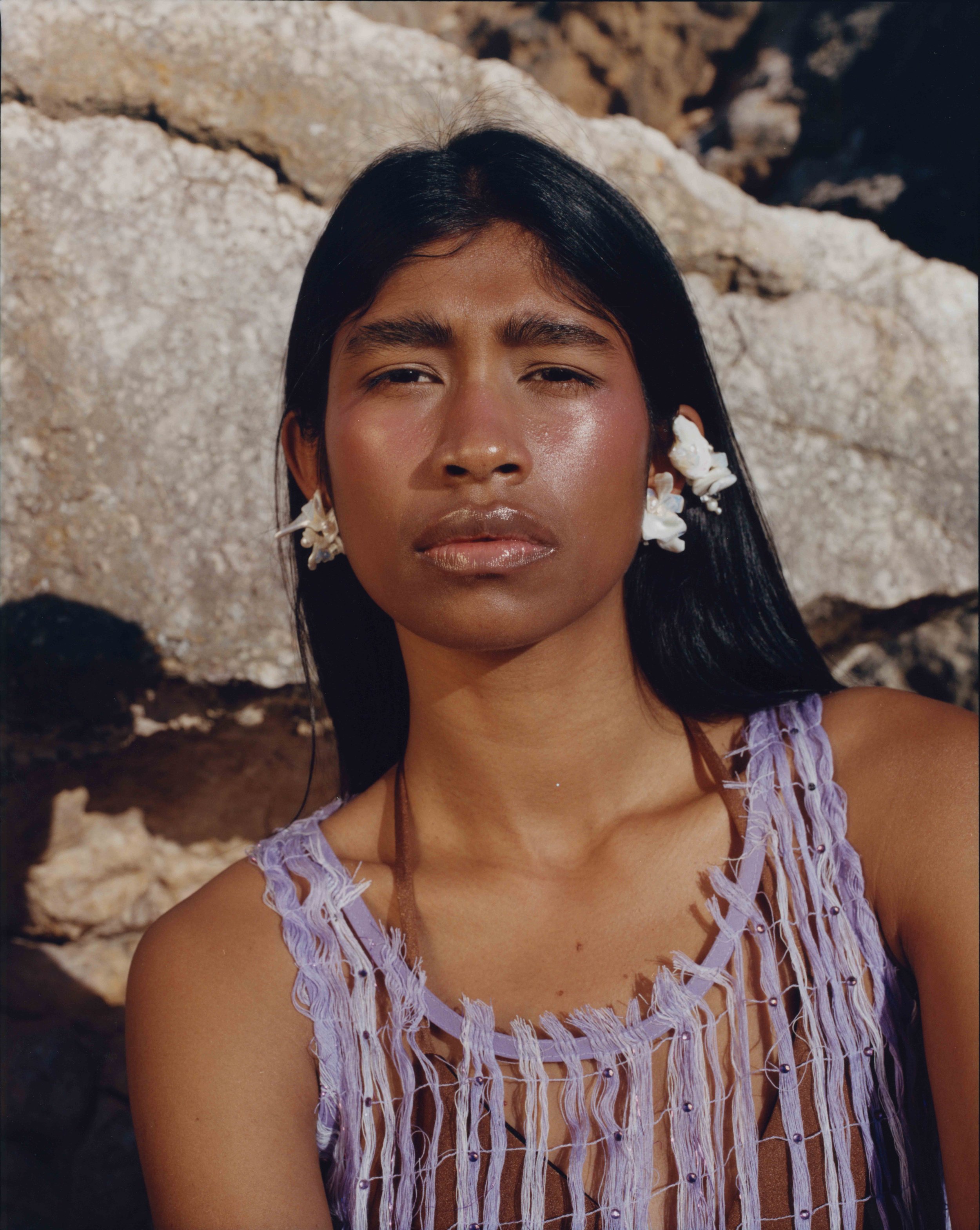 A close-up of a woman with long black hair, adorned with white shell earrings, wearing a purple fringed top against a rocky background.