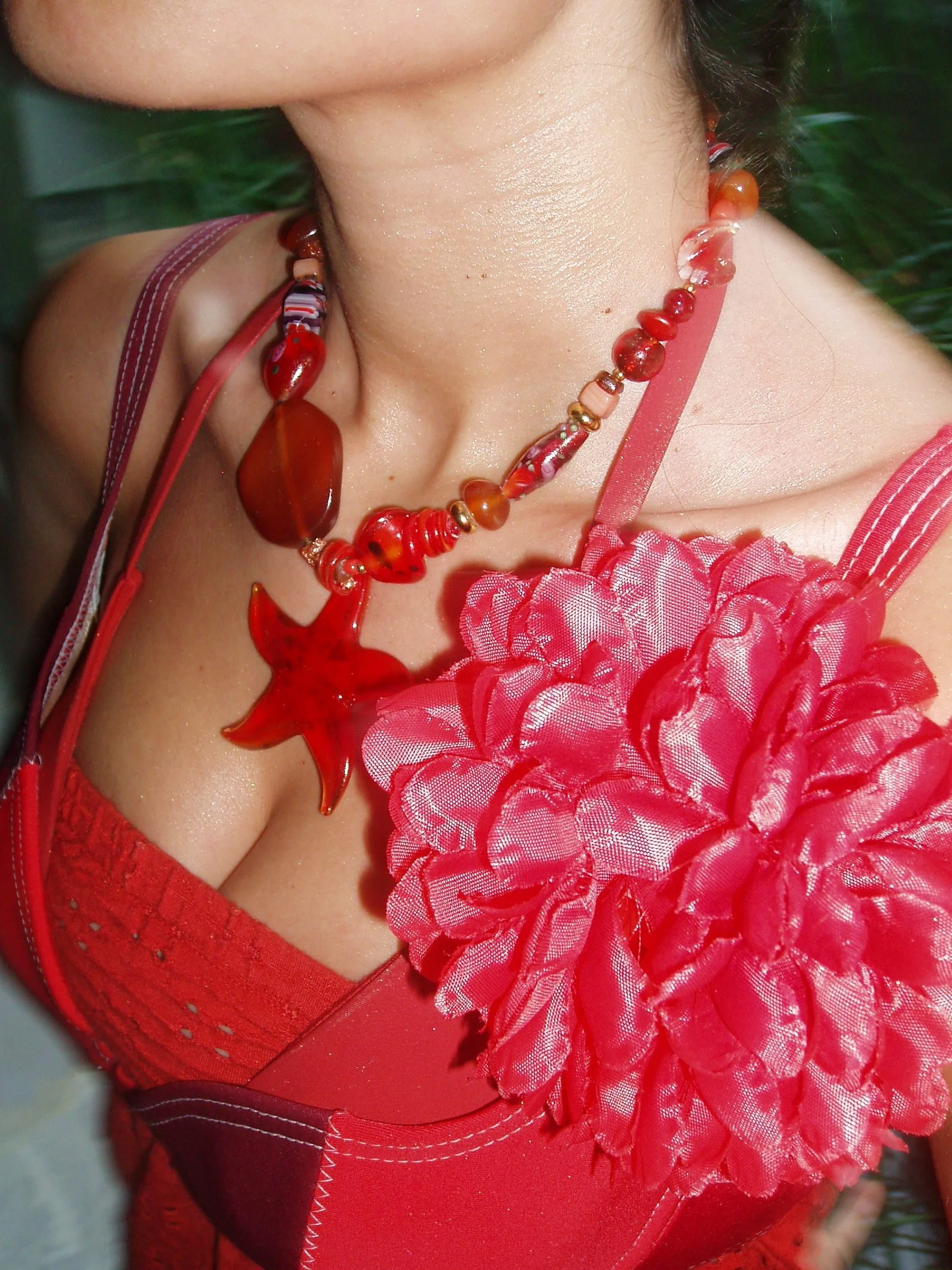 Close-up of a woman's neck, shoulder, and chest wearing a red dress with a large red fabric flower, and a necklace with red and orange beads and a star-shaped pendant.