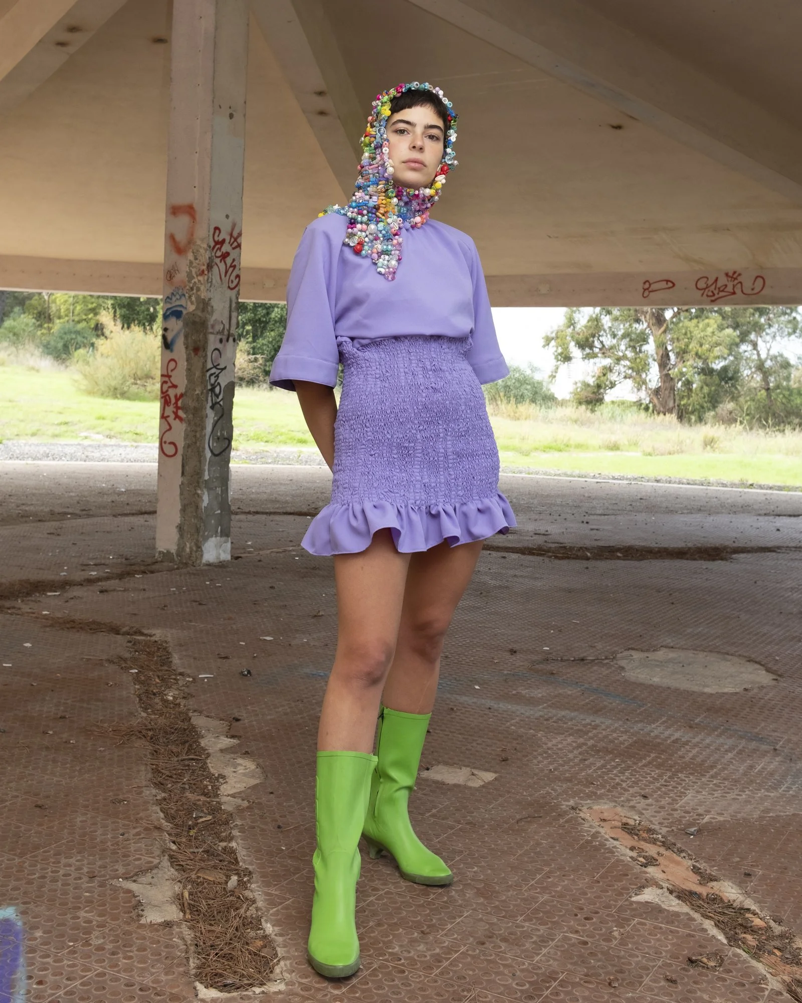 A woman with short dark hair standing under a concrete structure, wearing a purple ruffled dress, neon green boots, and a multicolored beaded hood.