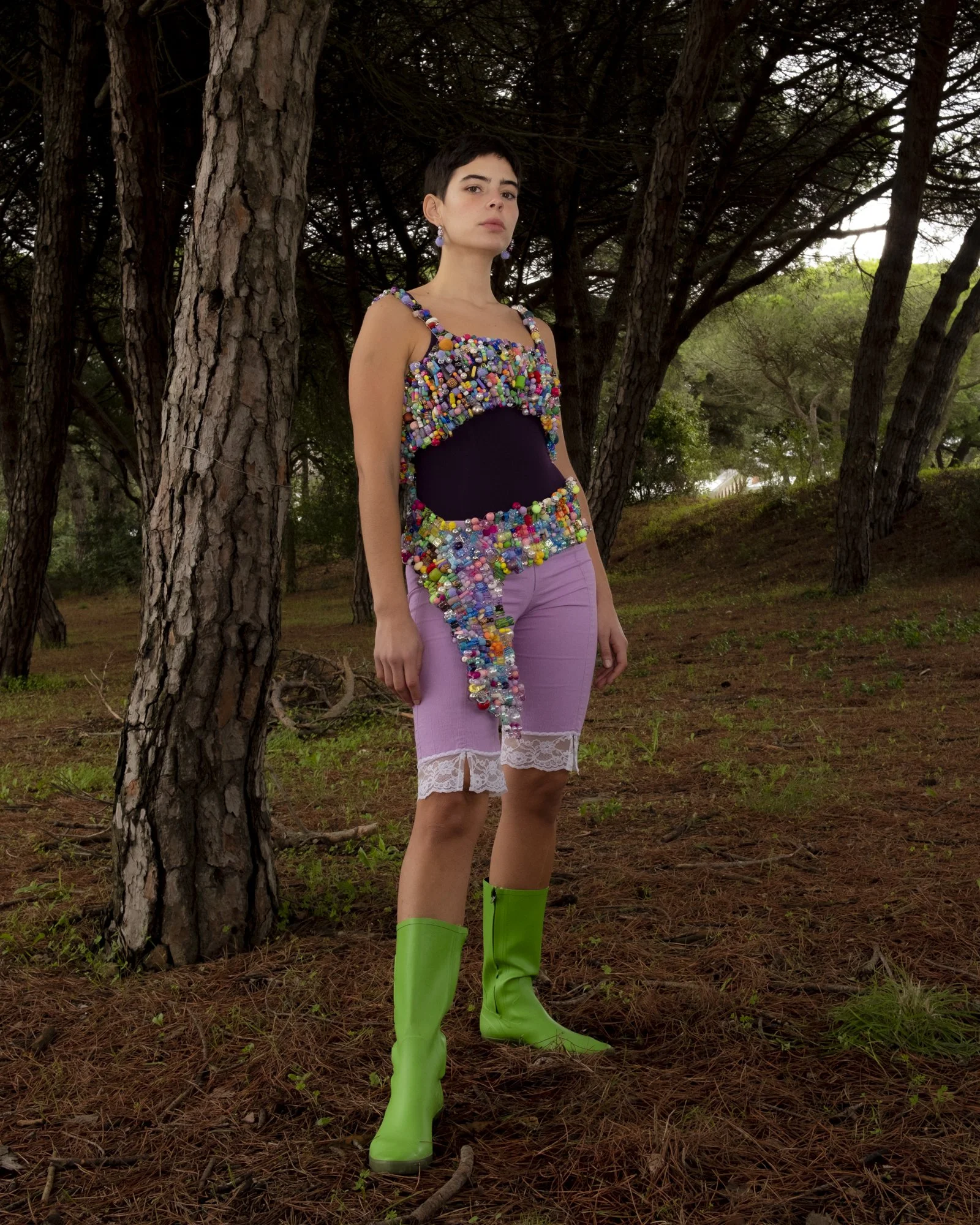 A woman standing in a wooded area wearing a colorful, beaded top, pink lace shorts, and bright green knee-high boots.