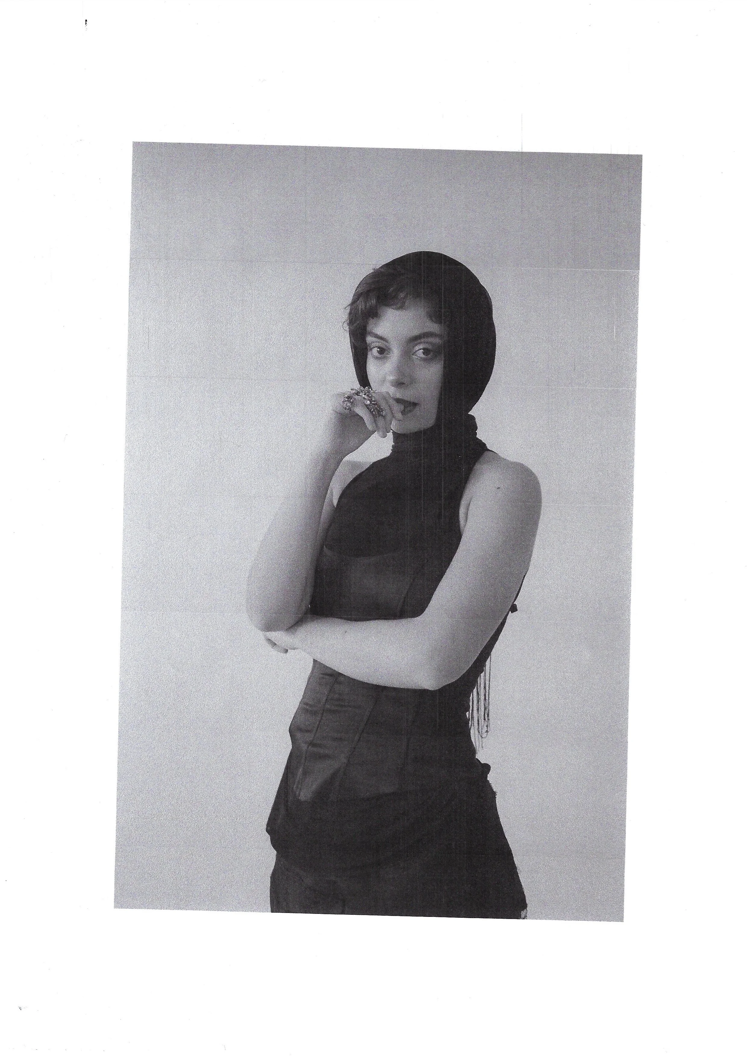 Black and white photo of a woman with short hair, wearing a dark sleeveless top and a dark headscarf, posing with her hand near her mouth and looking at the camera.
