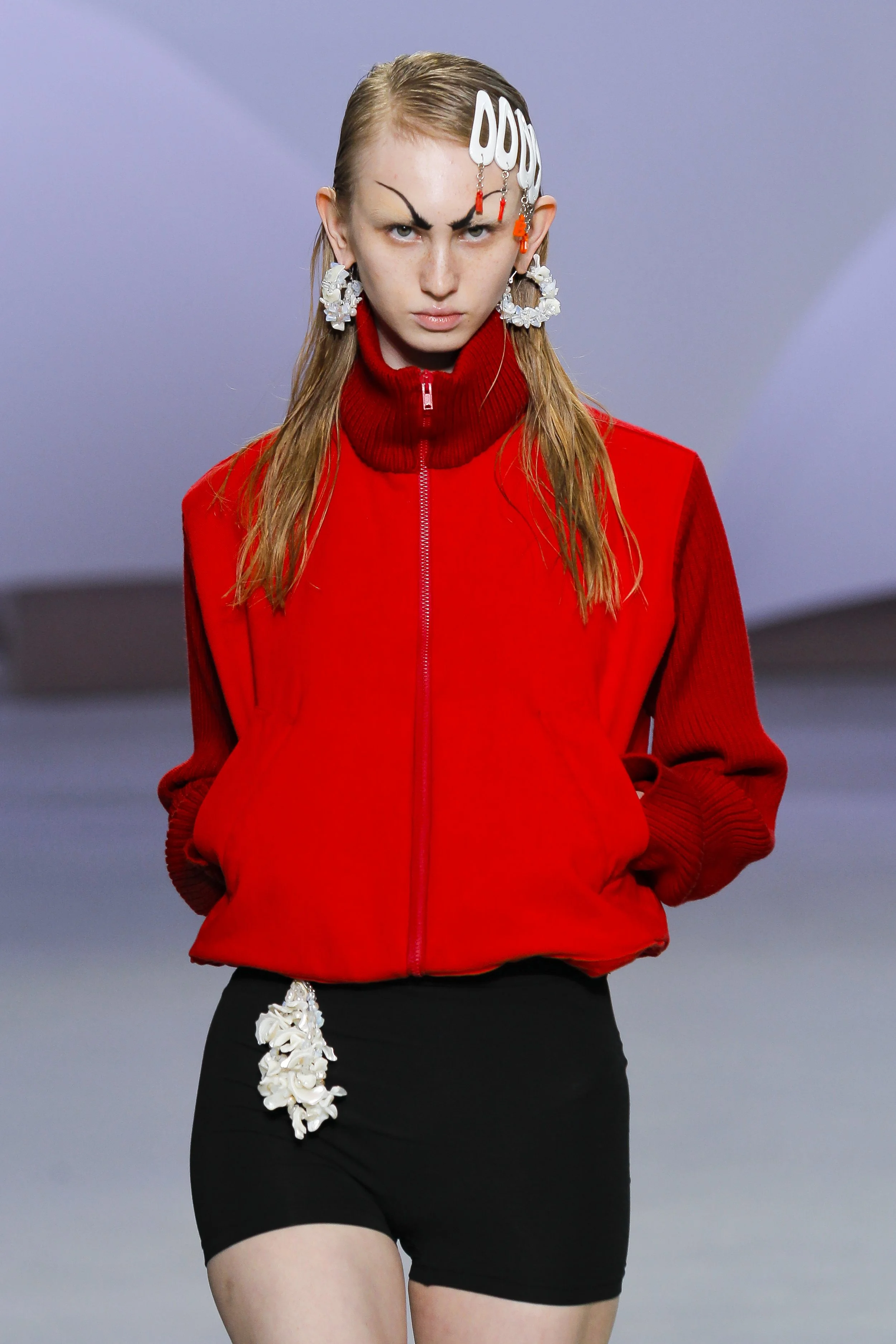 A female model walking on a runway, wearing a bright red jacket with a high collar, black shorts with a white decorative element hanging from the waistband, and large white earrings. Her hair is styled straight, and she has dramatic eyebrows.