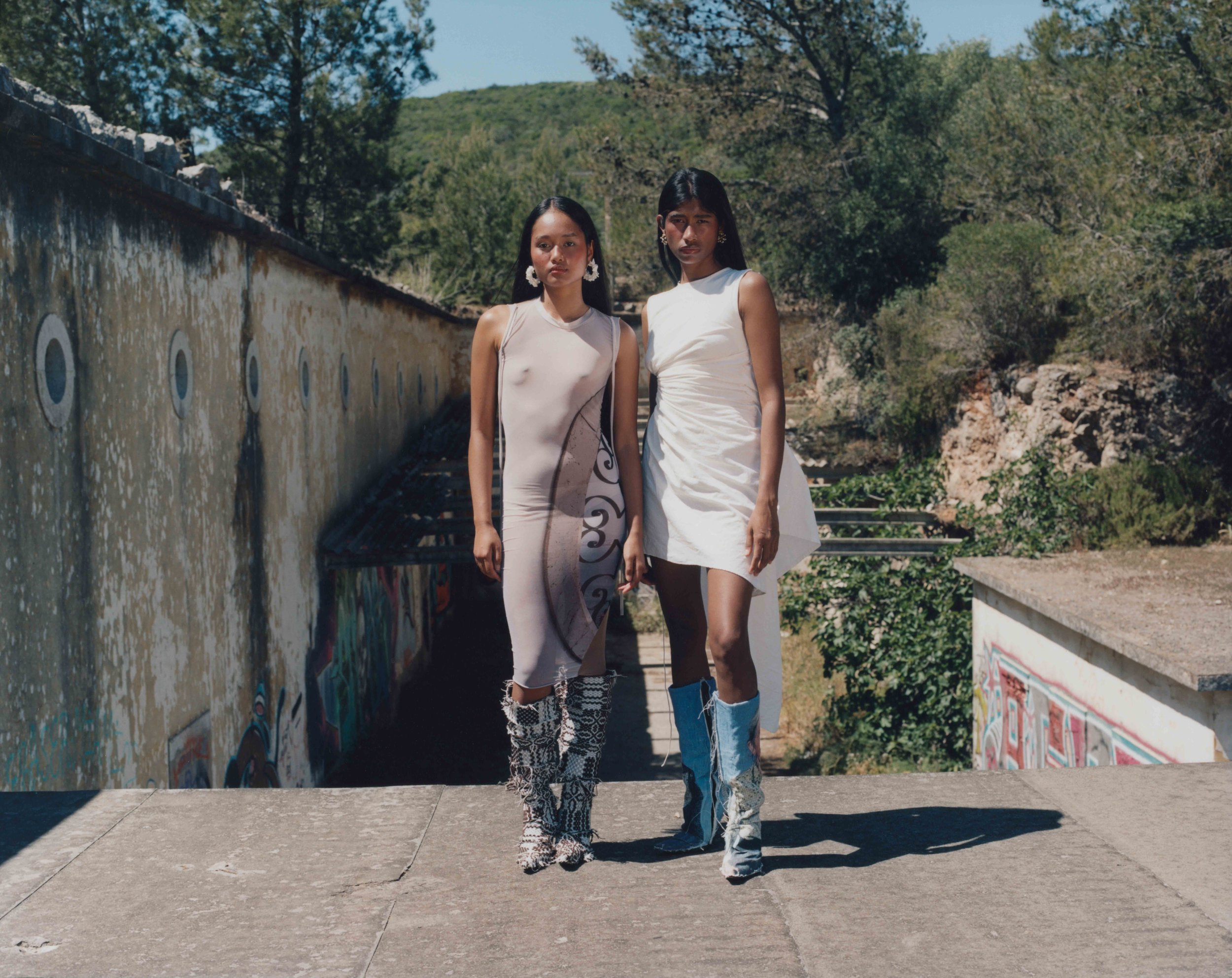 Two women standing outdoors on a concrete surface, with graffiti-covered walls, trees, and hills in the background. They wear fashionable dresses and bold boots, posing with serious expressions.