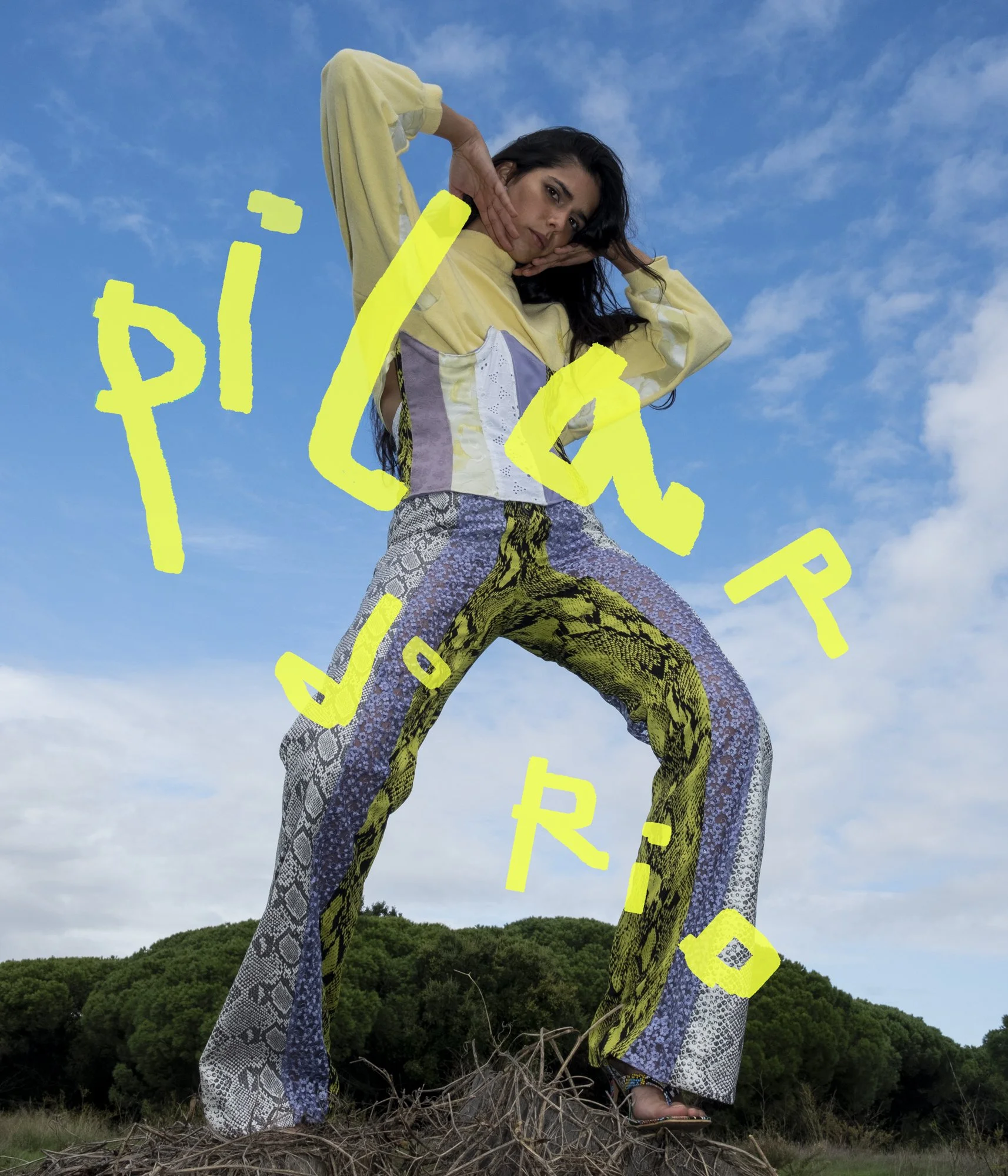 A woman with long dark hair wearing a yellow and patterned outfit stands outdoors on a pile of branches with a green, wooded hill and a blue sky with a few clouds in the background.
