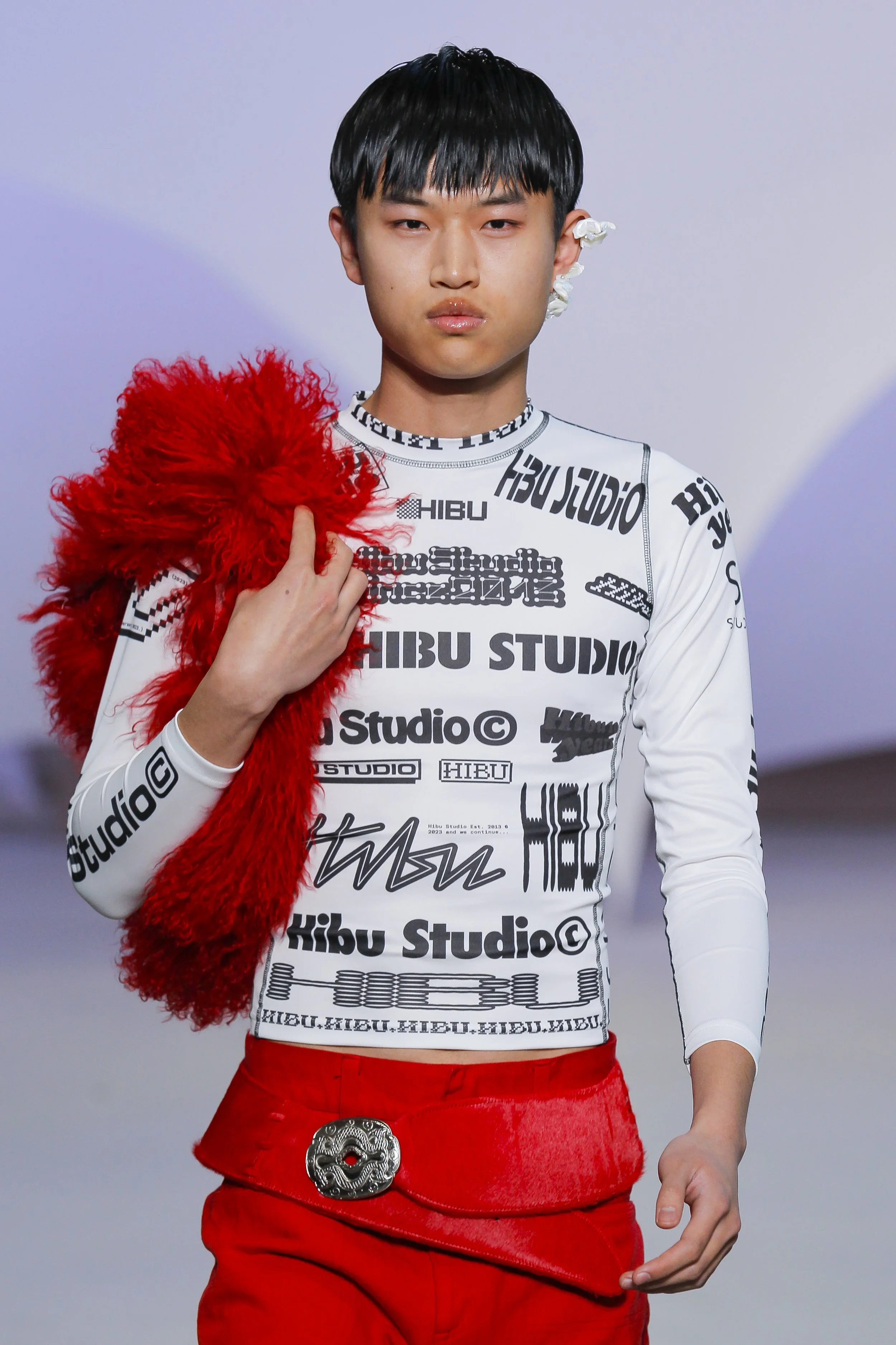 Male fashion model walking on runway wearing red pants with a large decorative belt, a white long-sleeve shirt covered in black text and patterns, holding a red furry accessory over his shoulder, with floral design earrings, a neutral expression, and