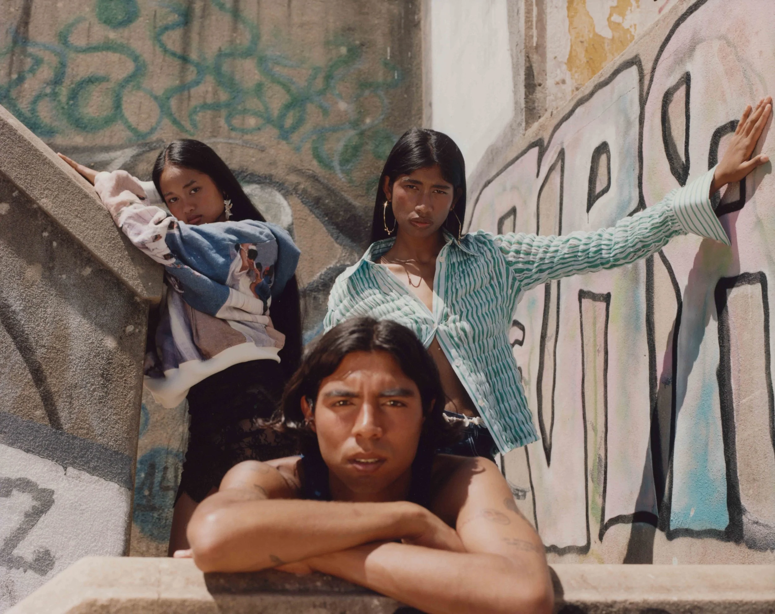 Three young people pose against graffiti-covered walls; one woman leans on the wall with her hand, another woman stands with one hand on the wall, and a man rests his chin on his crossed arms in front of them.