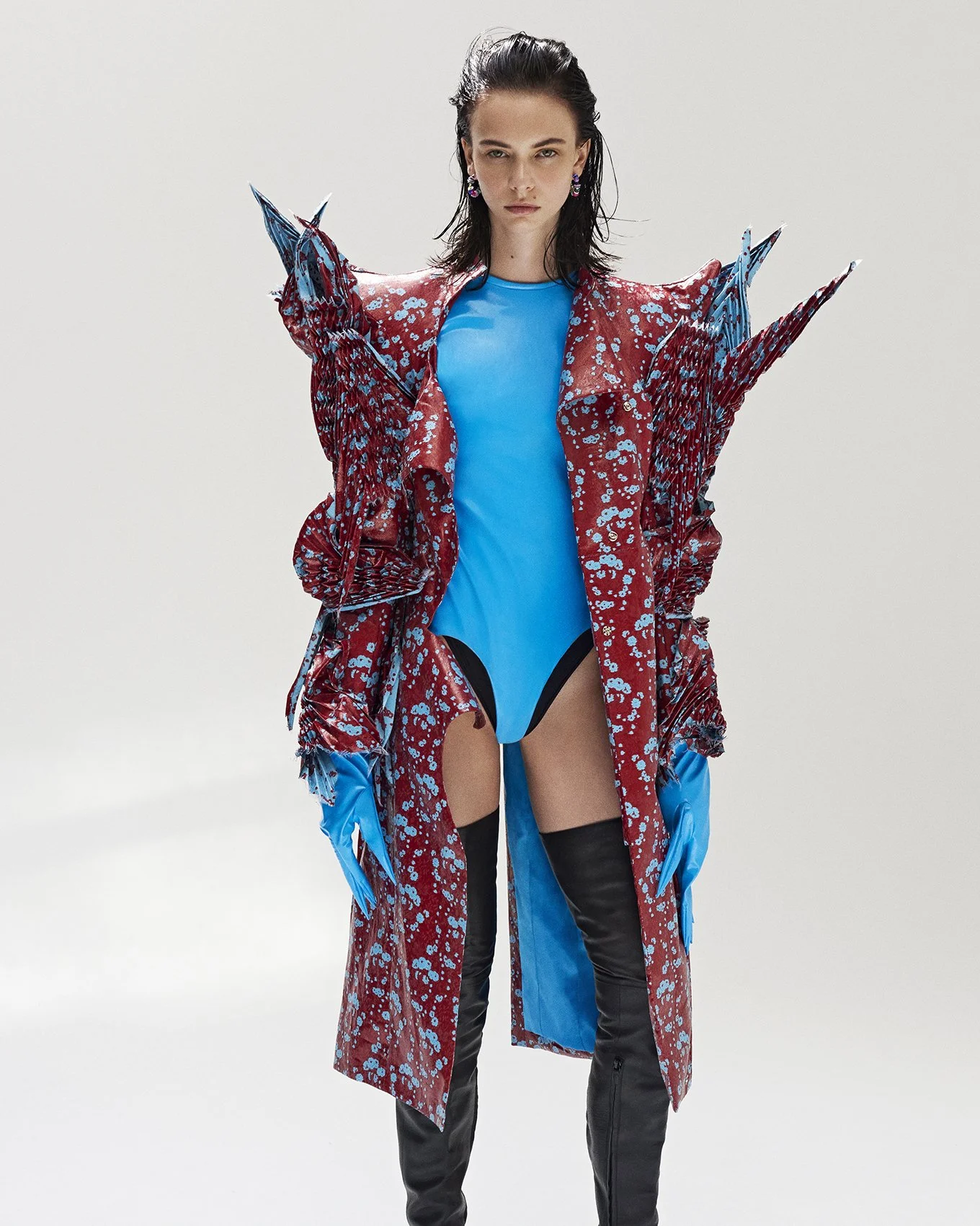 Model wearing avant-garde fashion with a red and blue textured coat, bright blue bodysuit, and thigh-high black boots, standing against a plain background.
