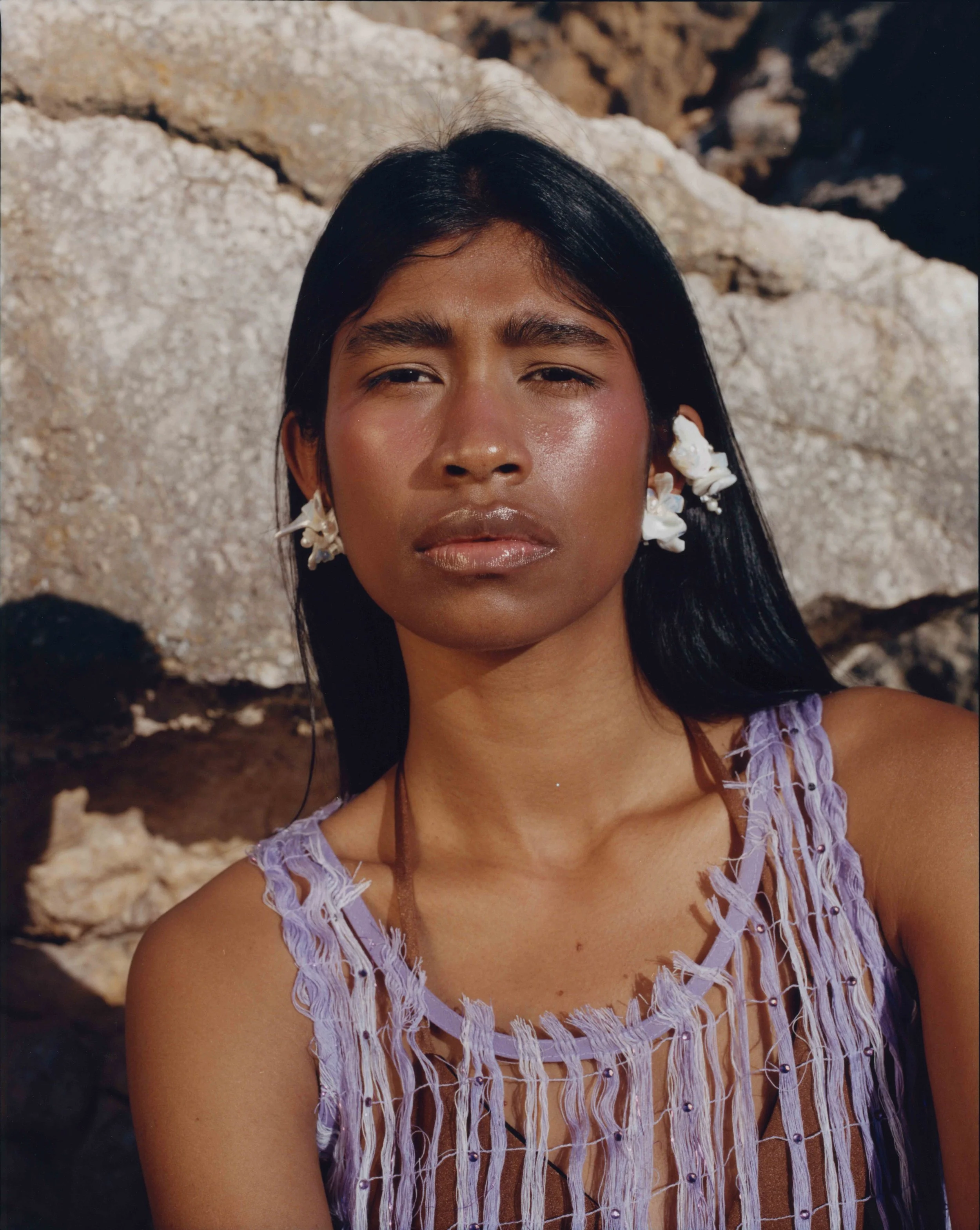 A woman with long black hair, wearing shell earrings and a purple top with fringe, is outdoors with rocks in the background. vogue editorial