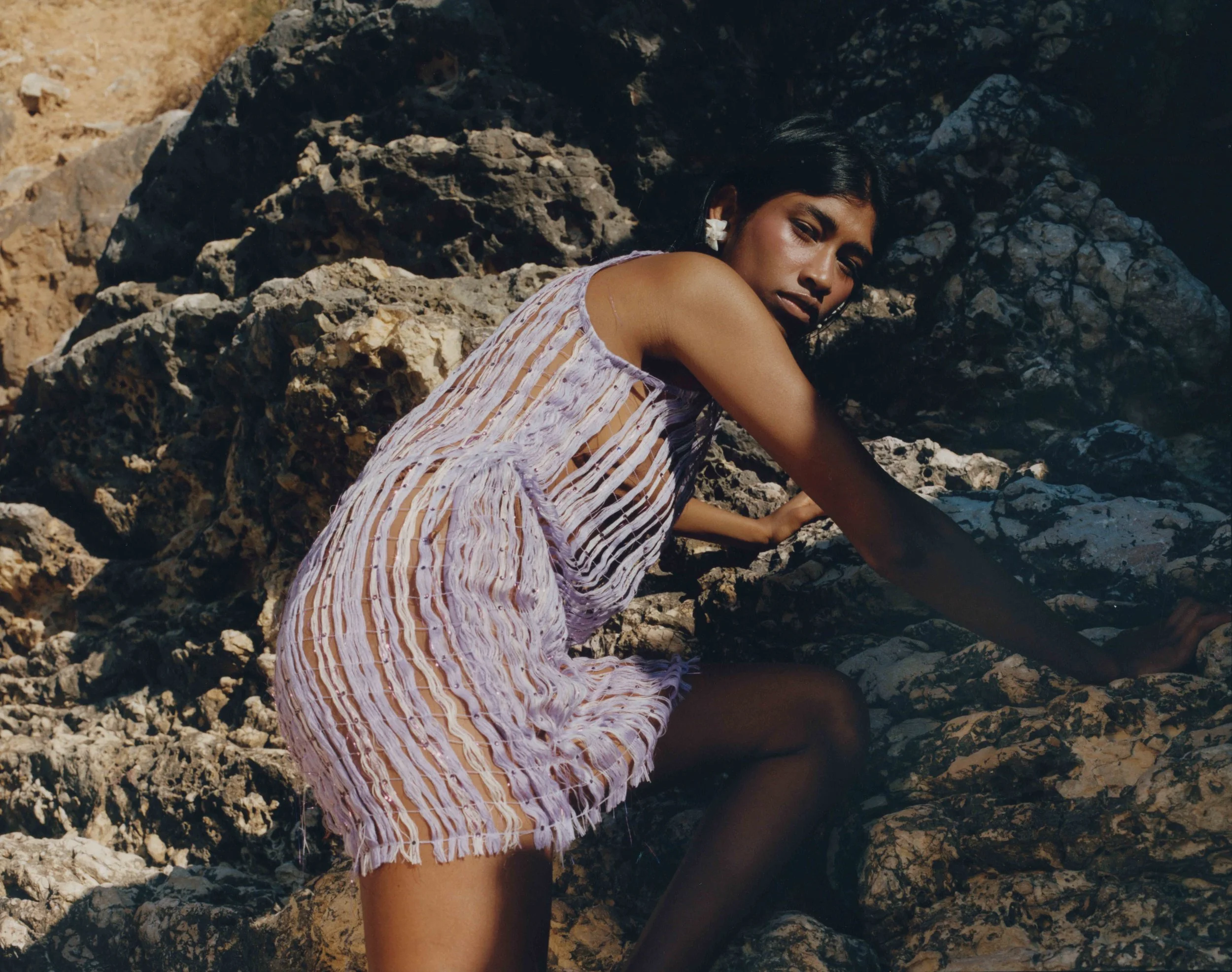 Woman with dark hair wearing a purple and white striped dress, climbing on rocks outdoors. vogue editorial