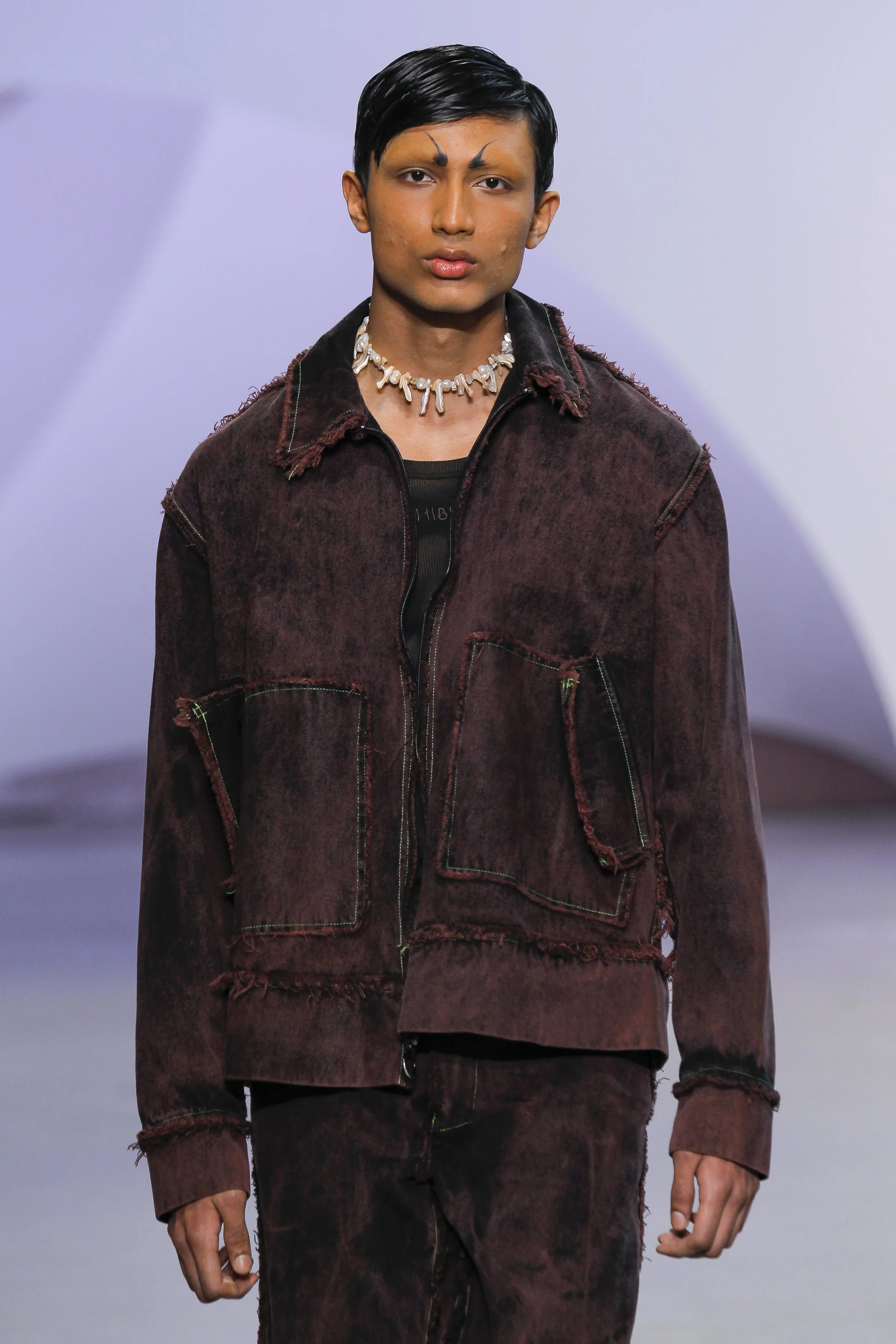 A male model walking on a runway, wearing a distressed, dark red jacket and matching pants, with a black shirt underneath, a seashell necklace, and short black hair.