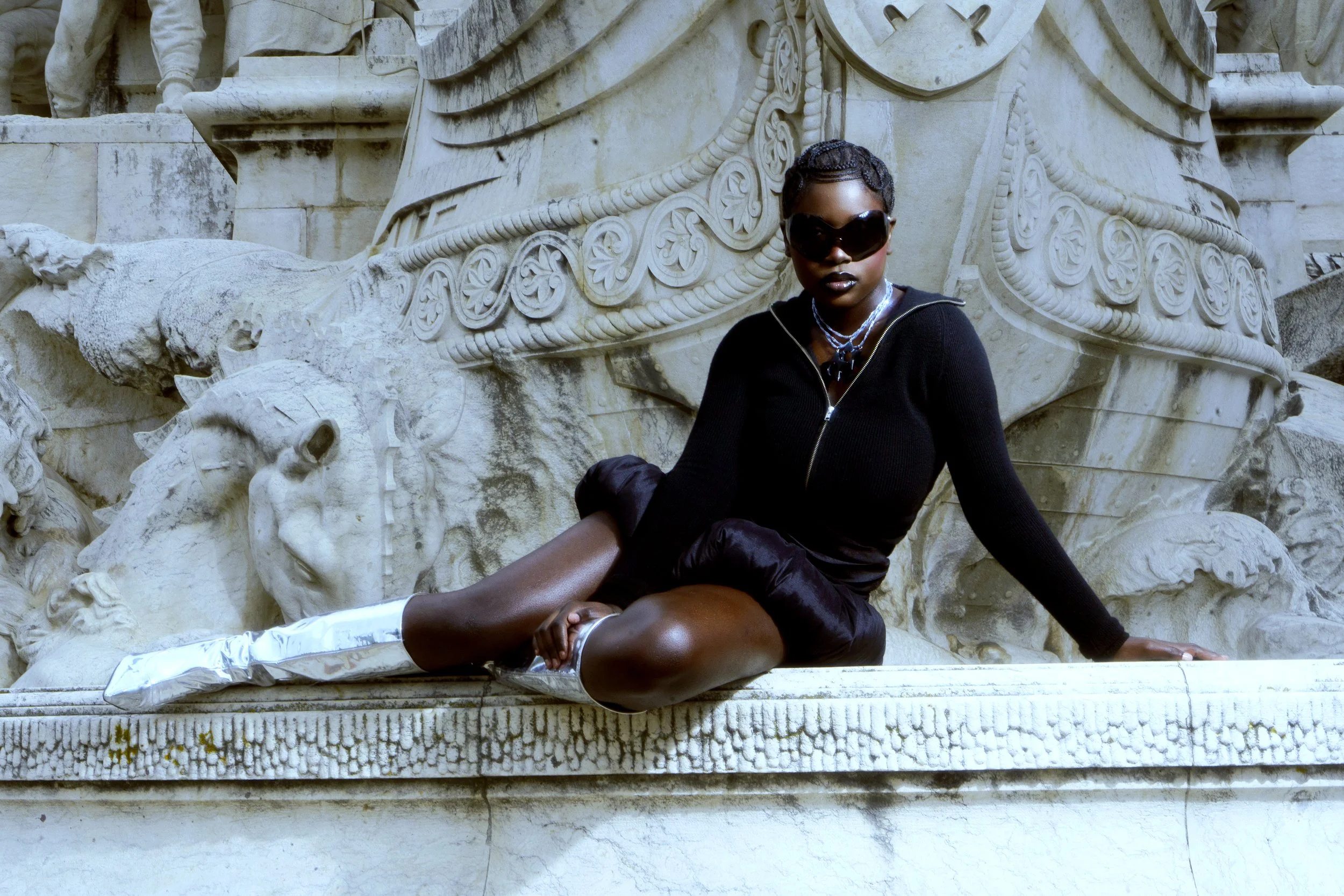 A woman with dark skin wearing sunglasses, a black zip-up jacket, black shorts, and white thigh-high boots sitting on a marble structure with carved details and sculptures.