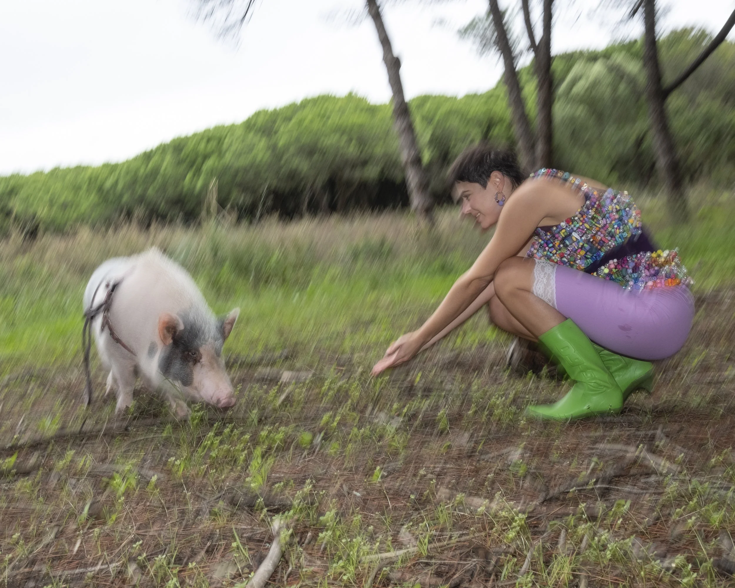 A woman crouching in a field, reaching out to a pig with black and white spots. The woman is wearing a colorful, beaded top, purple shorts with white lace, and bright green rain boots. The background features trees and greenery, with a cloudy sky.
