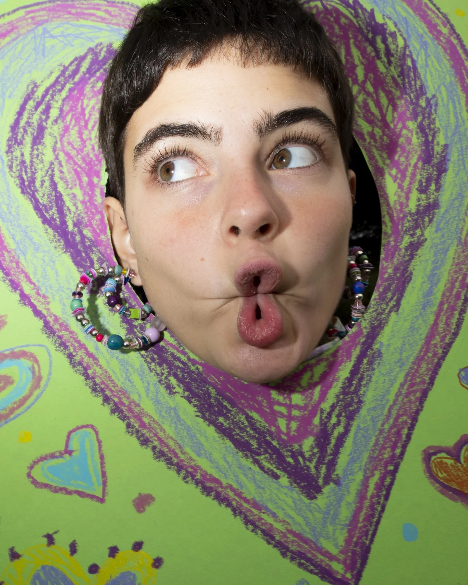 A young person with short dark hair and brown eyes makes a funny face by pursing their lips with a small hole in the middle, looking to the side. They are wearing colorful beaded earrings and are positioned behind a cutout of a green and purple heart