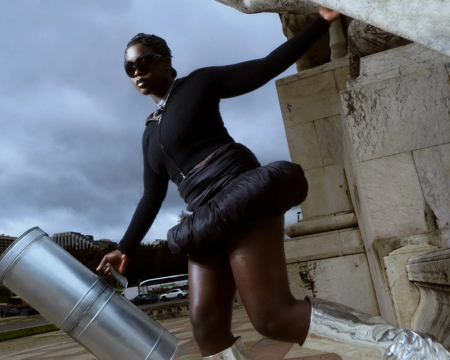 A woman wearing sunglasses, a black zip-up top, black shorts, and metallic knee-high boots is kneeling on one knee beside stone architecture outdoors during cloudy weather. She holds a silver container, possibly a trash can, in her right hand.