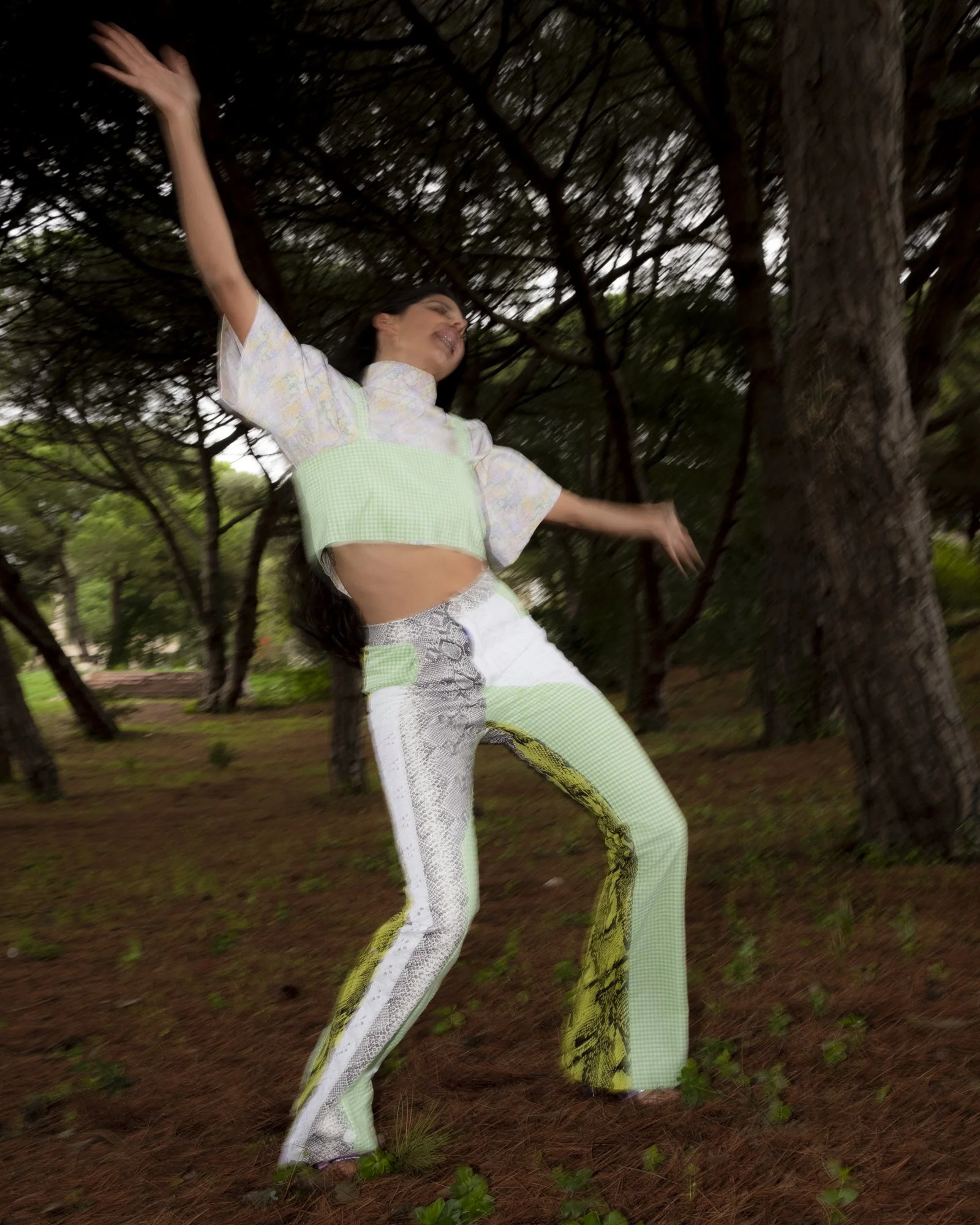 A woman dancing joyfully in a forested area with tall trees and pine needles on the ground, wearing a stylish matching outfit that includes patterned pants and a cropped top.