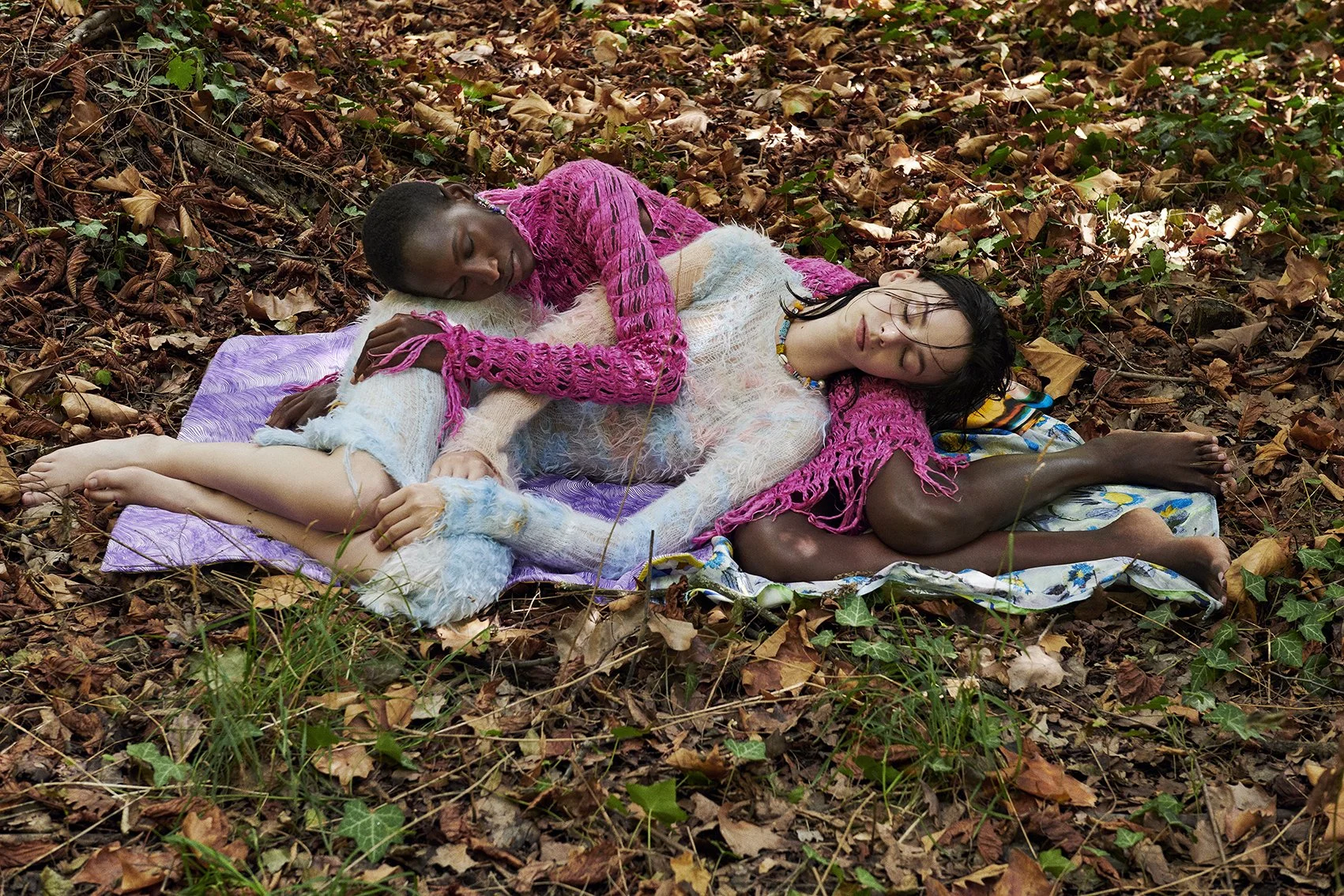 Two women lying on a blanket on the forest floor, surrounded by fallen leaves and greenery, embracing each other peacefully with their eyes closed.