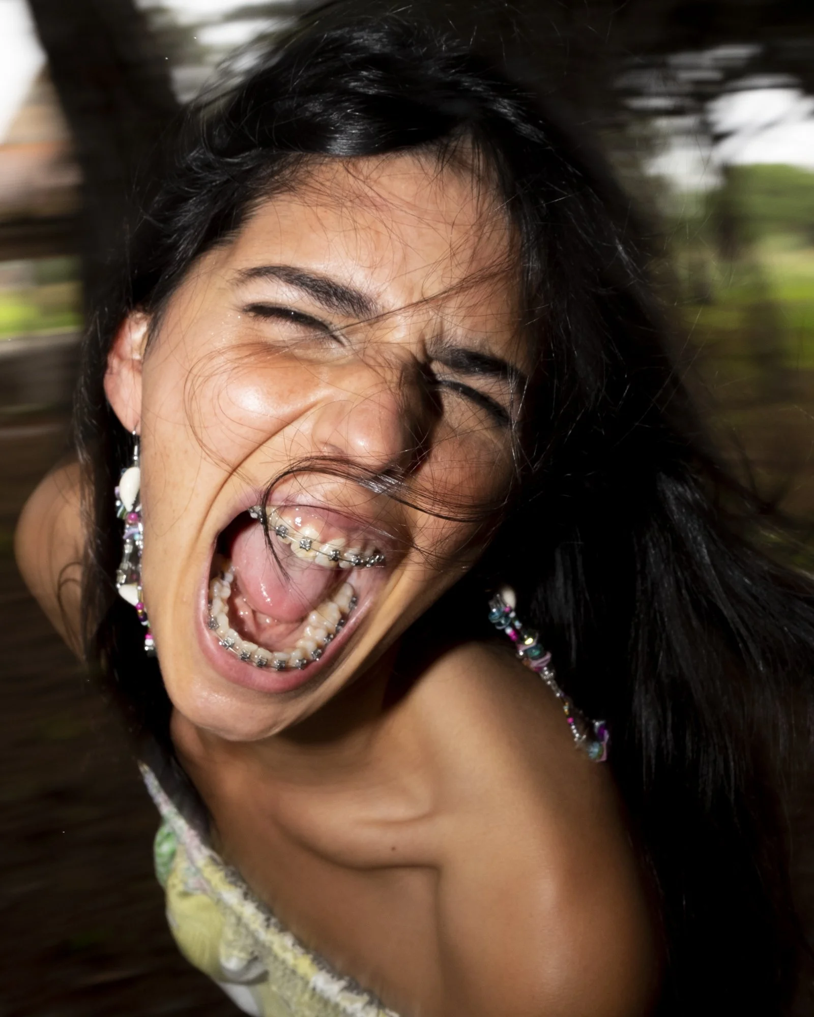 Close-up photo of a woman with braces and dark hair, laughing or shouting with her mouth wide open, showing her teeth and tongue, and wind-blown hair.