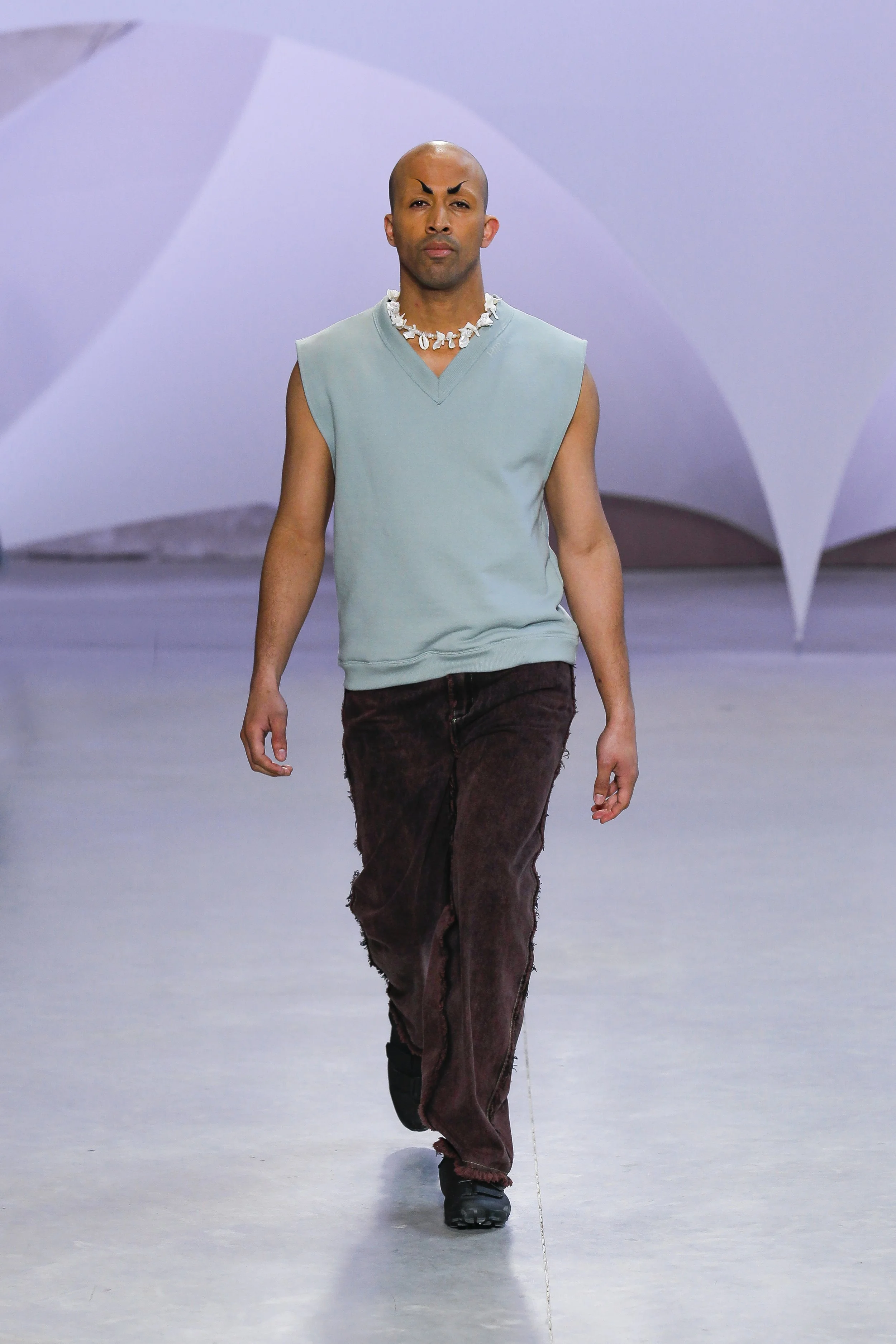 A man with a shaved head and bold black eyebrows walks on a runway, wearing a sleeveless light blue top, dark brown pants, black shoes, and a shell necklace, with abstract background.