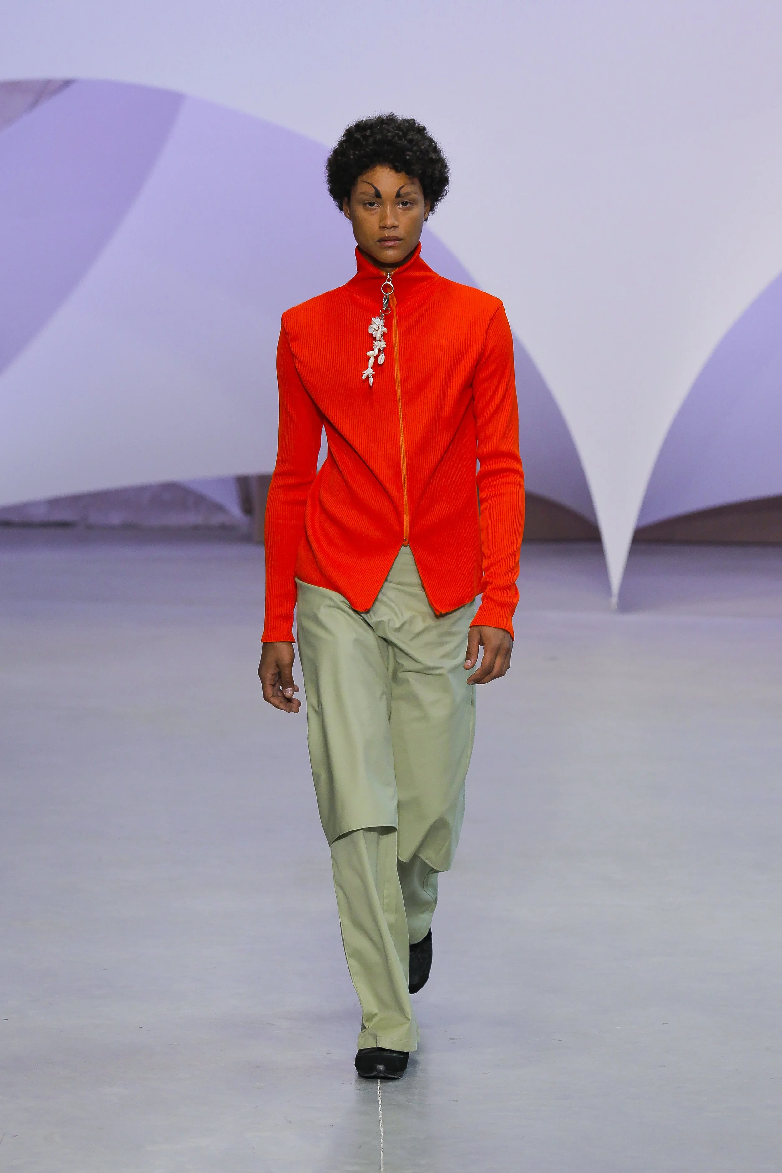 Fashion model walking on runway in orange zip-up jacket and beige pants with large shades of gray and purple in background.