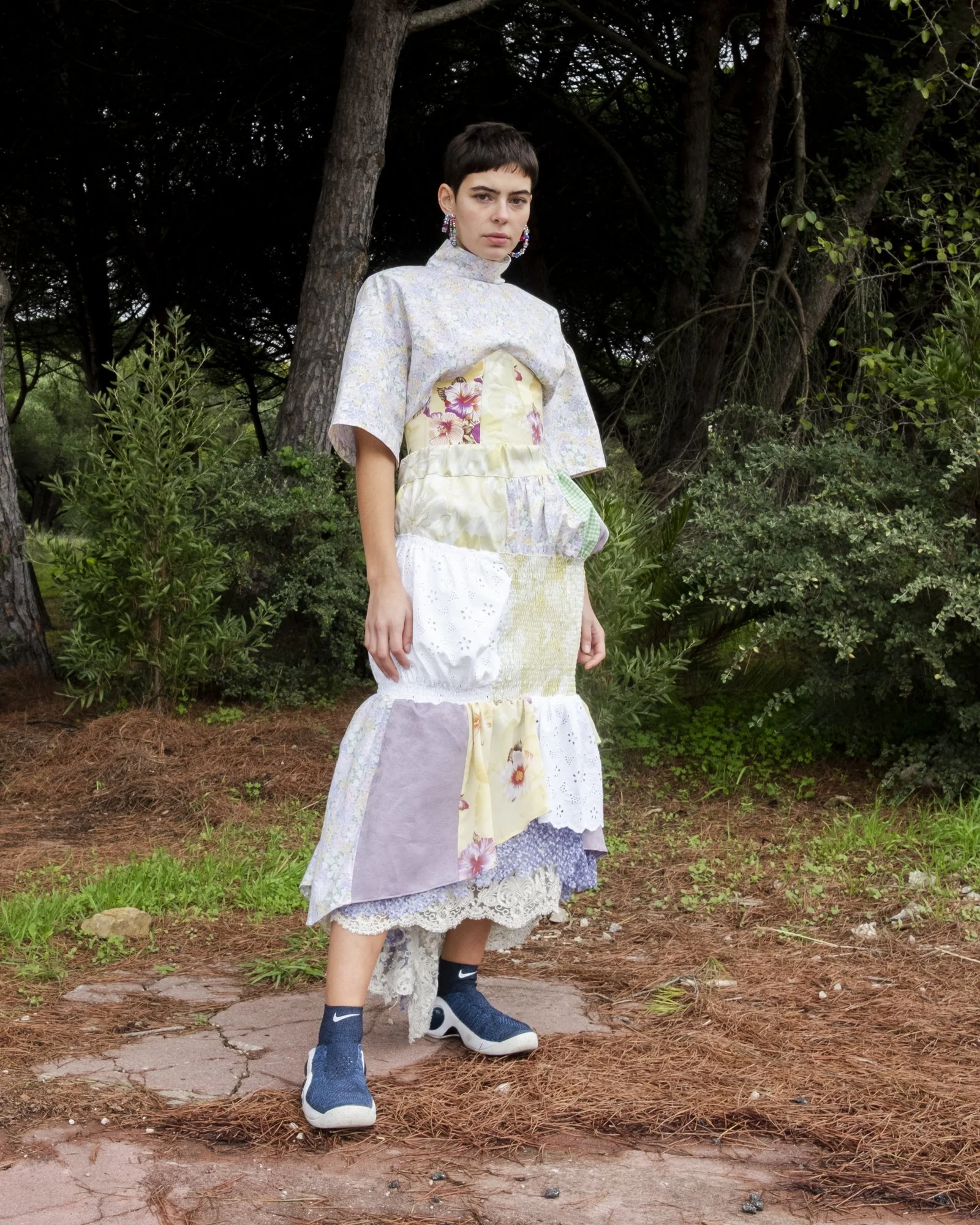 A woman wearing a patchwork dress with floral, lace, and fabric patterns stands outdoors in a wooded area.