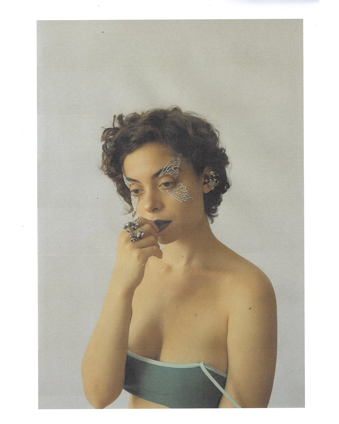 A woman with short, curly hair and dark lipstick, wearing rhinestone butterfly face stickers, multiple rings, and earrings, posing with her hand near her chin in front of a plain white background.