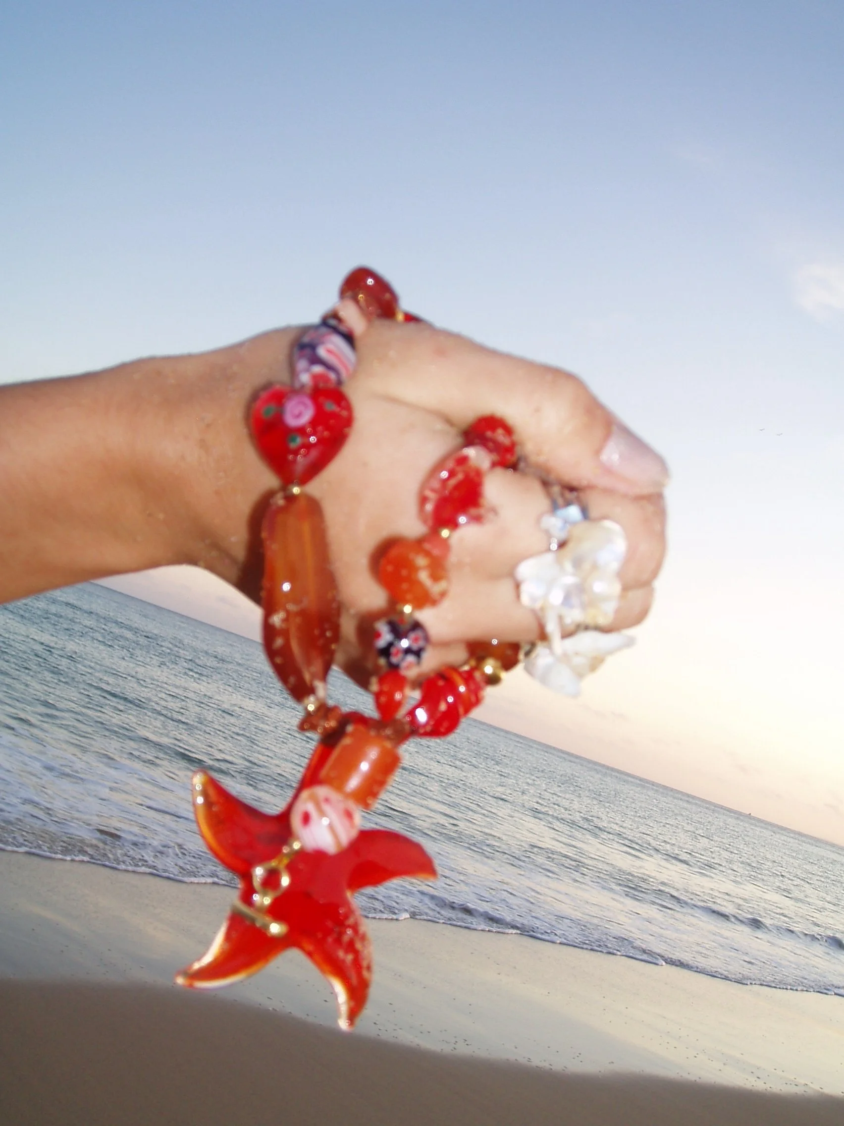 A person wearing a colorful bracelet is holding a coral-shaped object in the foreground while the ocean and sky are visible in the background.