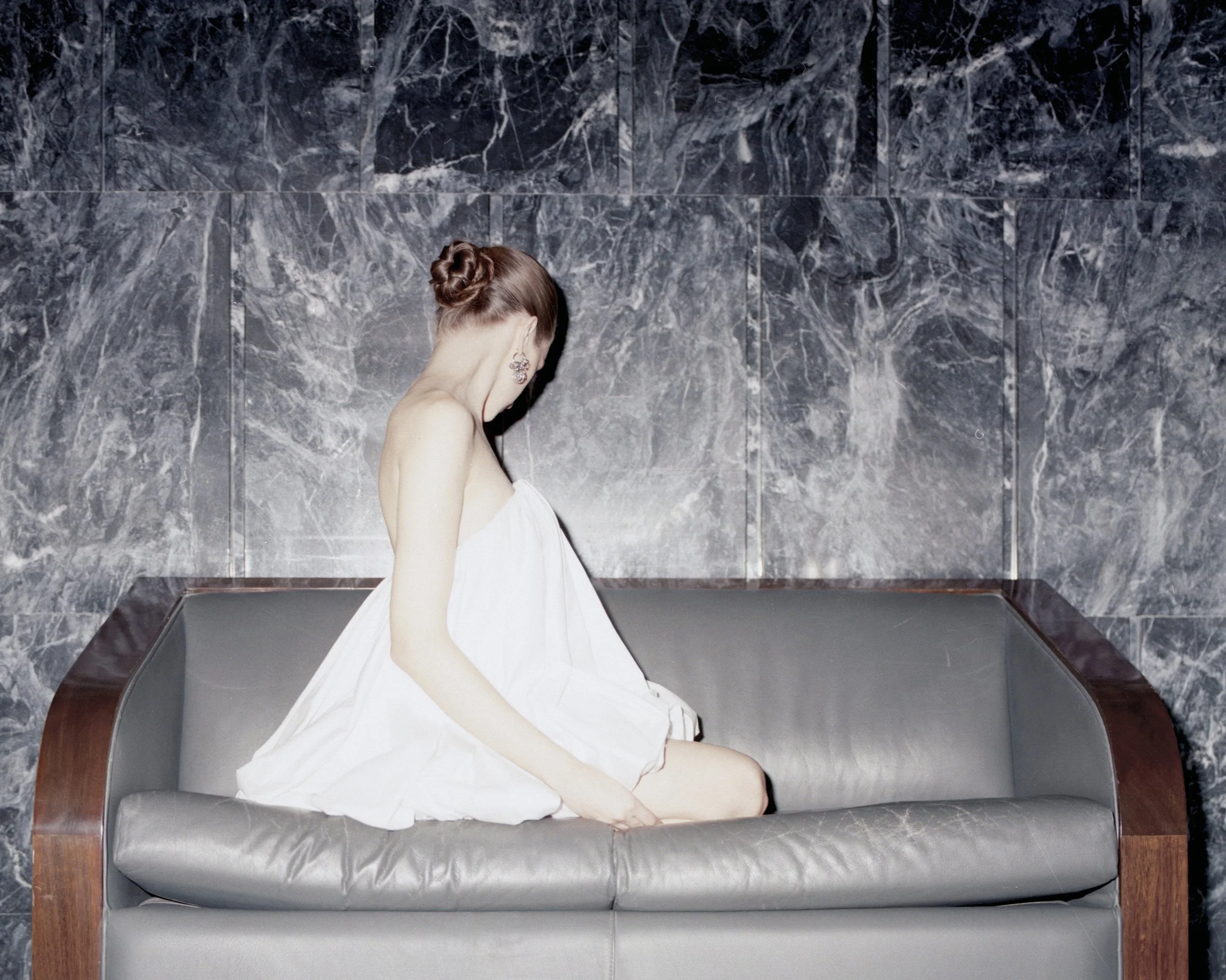 A woman in a white dress sitting on a gray leather sofa with a marble wall background.