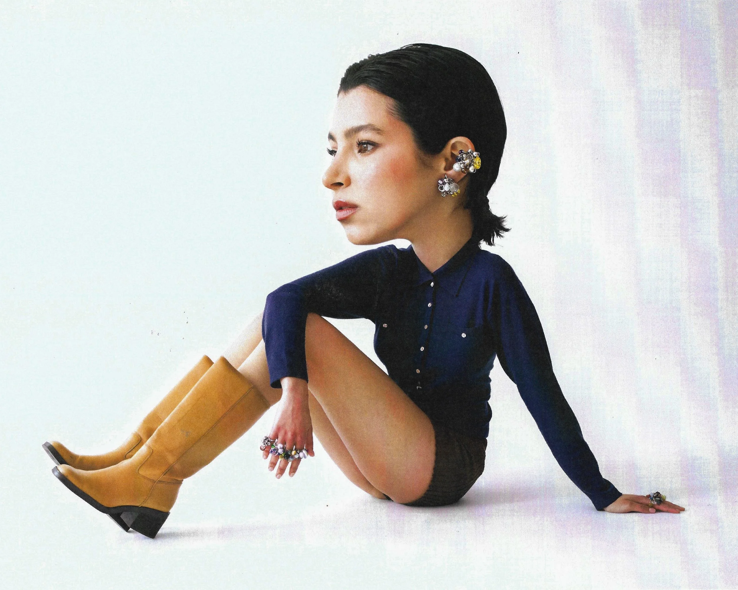 A woman with short dark hair, wearing a navy blue shirt, black shorts, and yellow boots, is sitting on the floor with one arm supporting her and the other resting on her knee. She is accessorized with multiple rings and earrings.