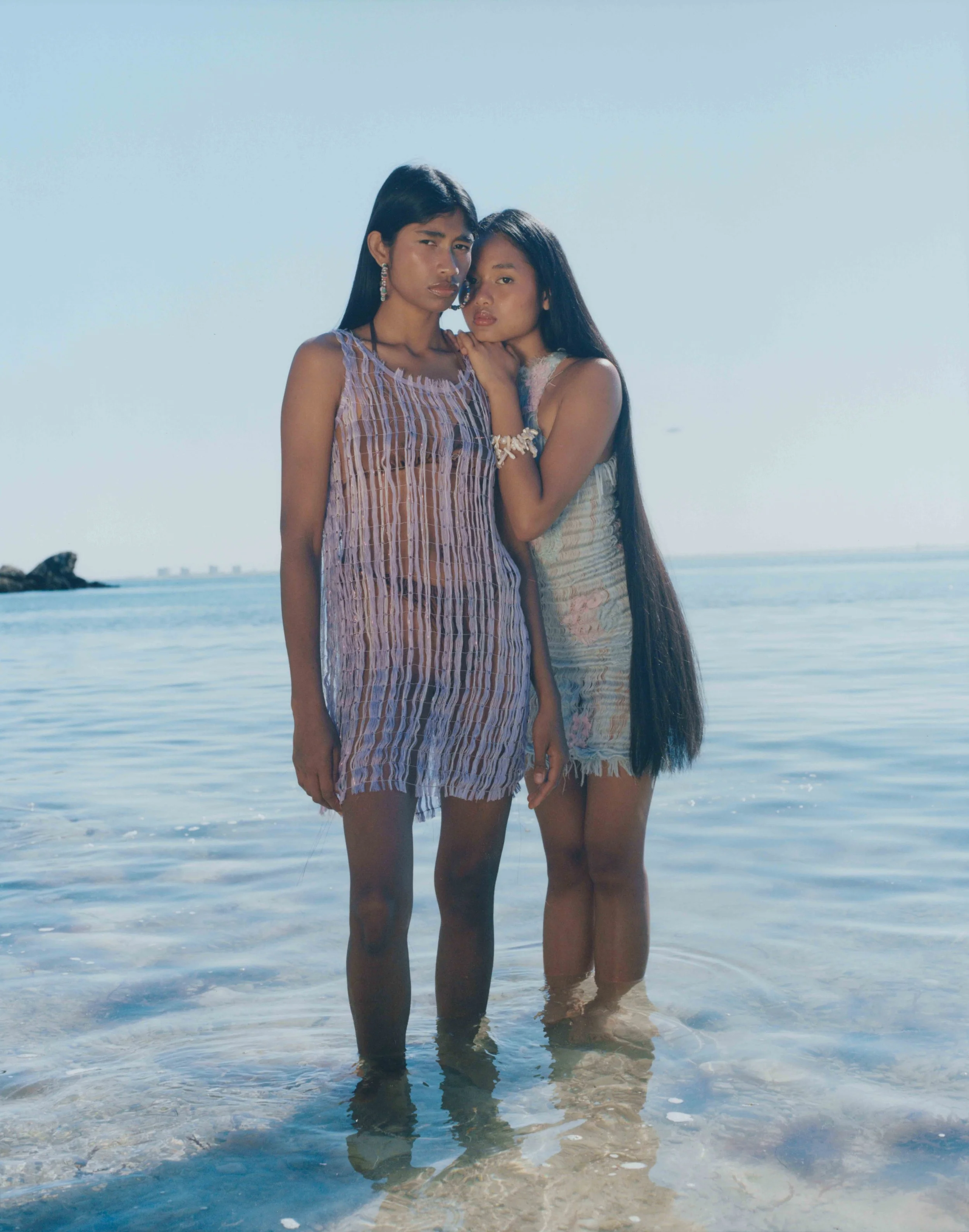 Two women with long dark hair standing in shallow ocean water, wearing light-colored dresses, during daytime with clear sky.