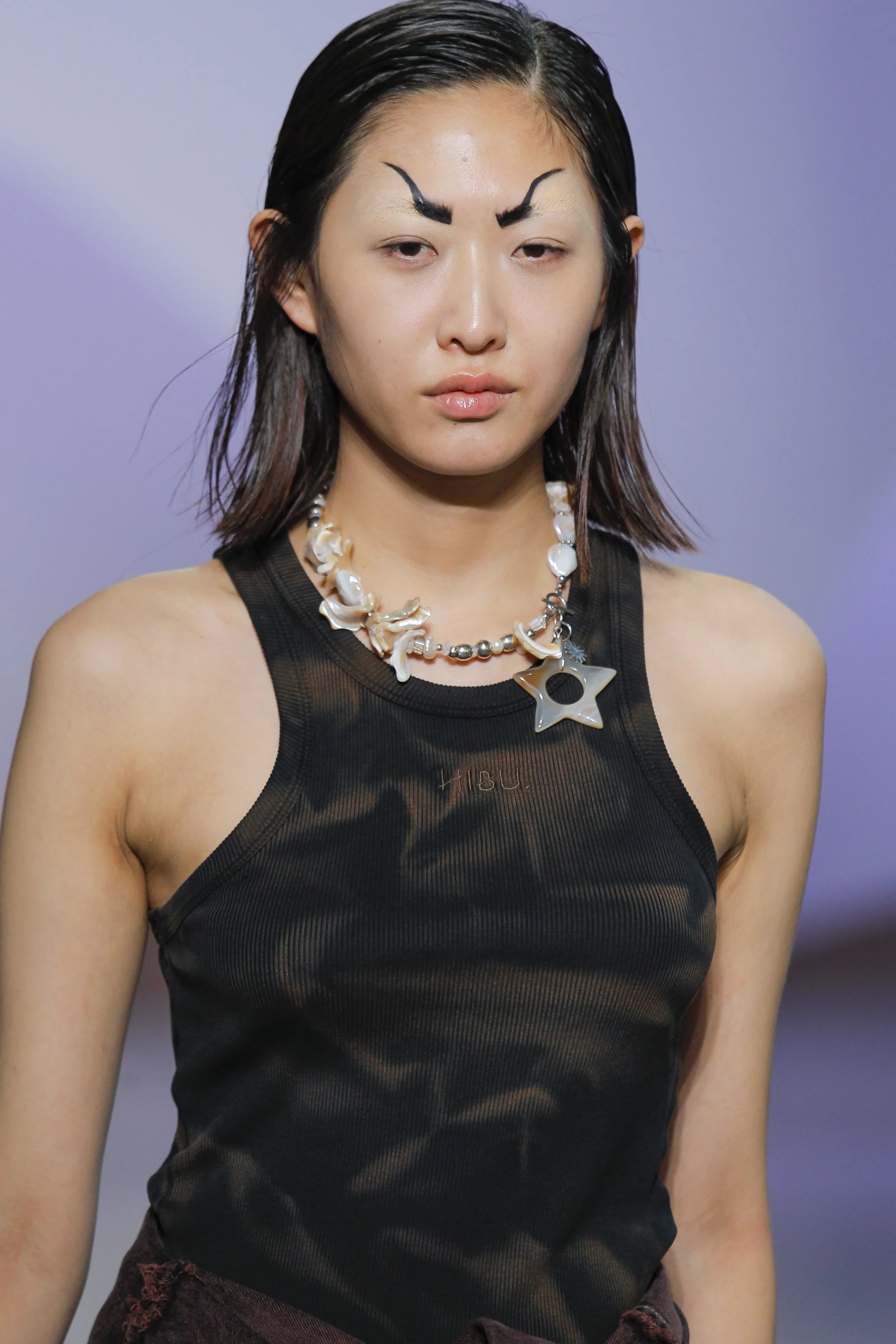 A young woman with dark, slicked-back hair wearing a black mesh tank top with the word 'HUB' on it. She has strange facial makeup that resembles black eyebrows painted above her natural eyebrows and no makeup around eyes, on a purple background.