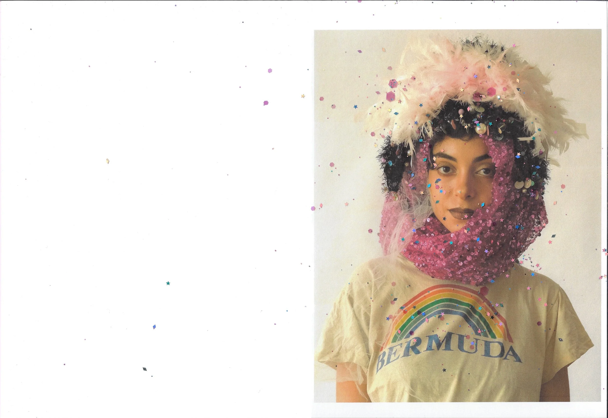 A person wearing a beige T-shirt with a rainbow and the word 'BERMUDA' on it, adorned with colorful sequins, feathers, and fabric, with a headpiece decorated with feathers, beads, and embellishments, surrounded by falling glitter.