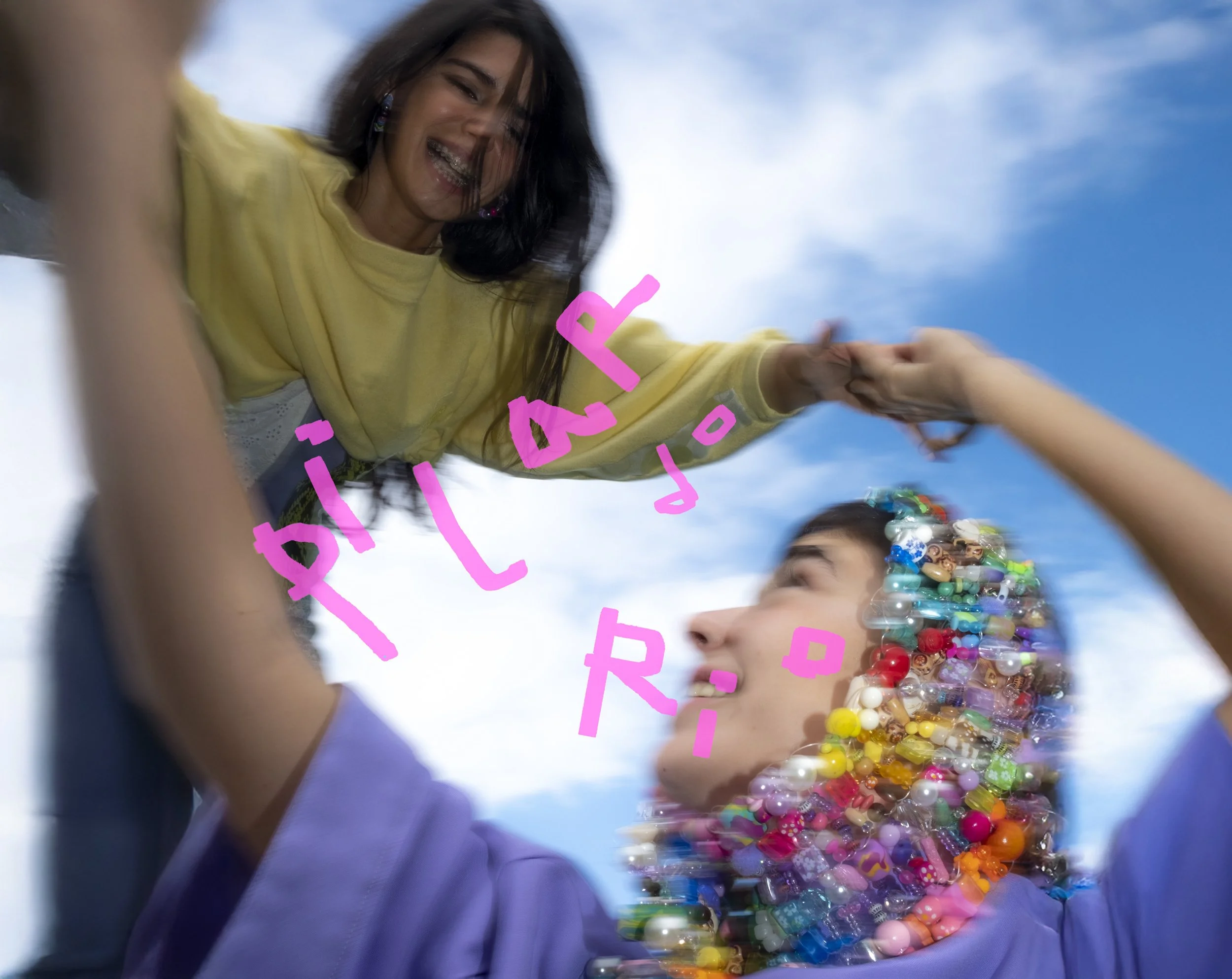Young girl smiling and reaching out to hold hands with someone, wearing a yellow shirt and colorful beaded jewelry, against a cloudy blue sky.