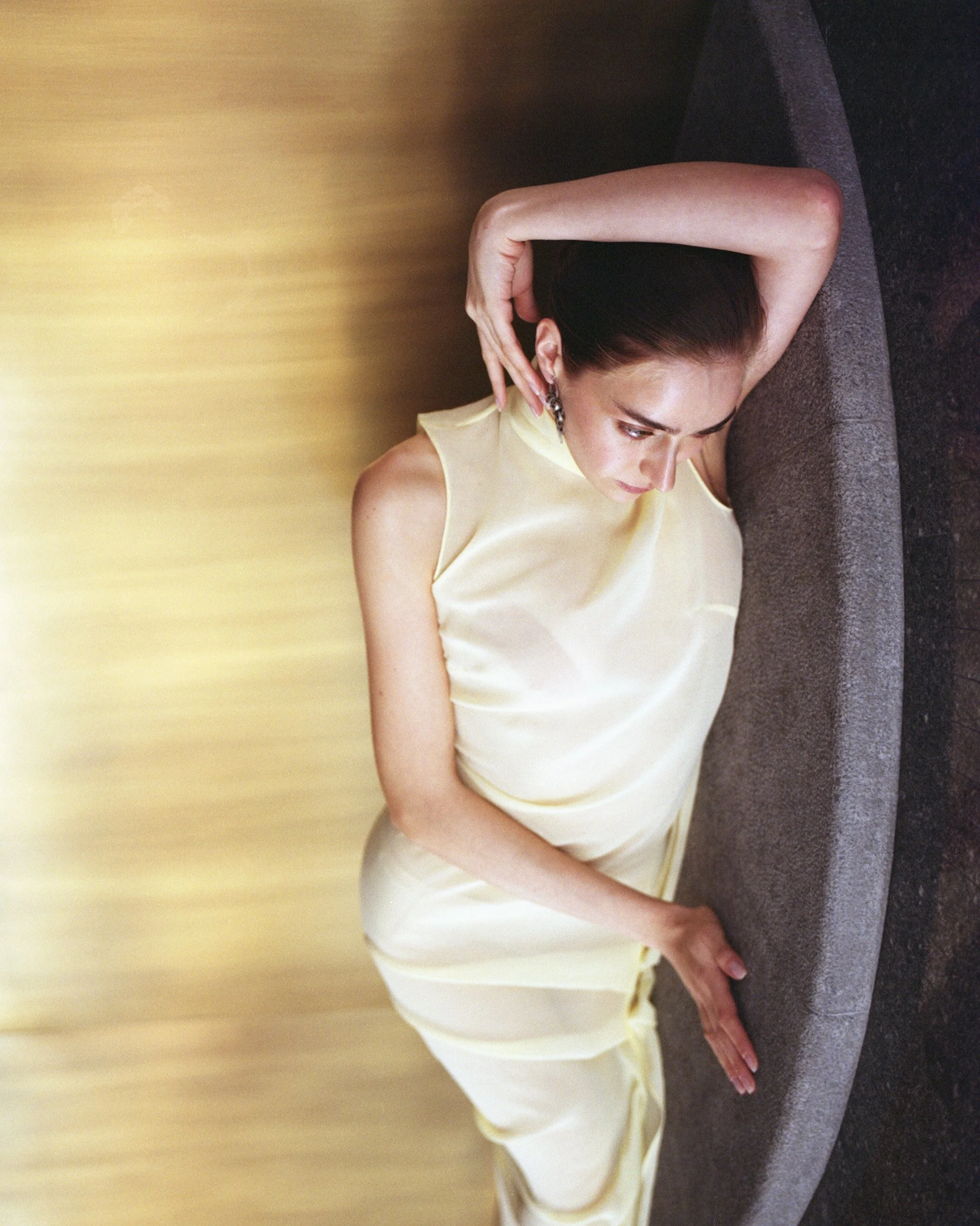 A woman in a light yellow satin dress lying on a curved gray sofa with her arm resting on her head and looking down.