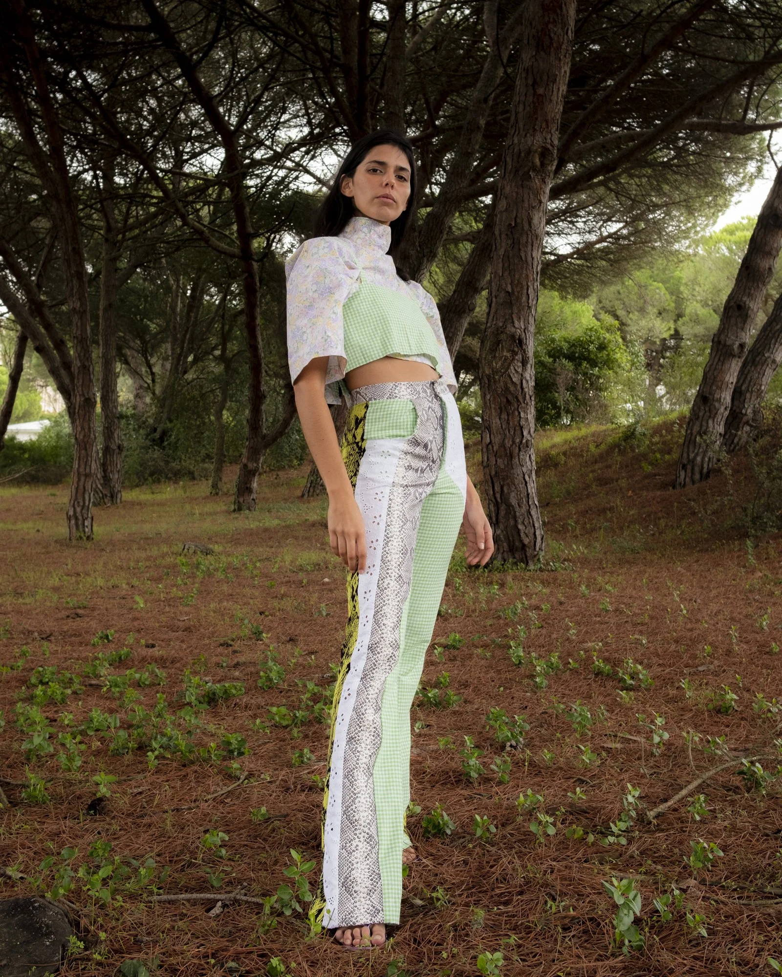 A woman standing in a forest, wearing a patchwork outfit combining different patterns and colors, with tall trees and earthy ground in the background.