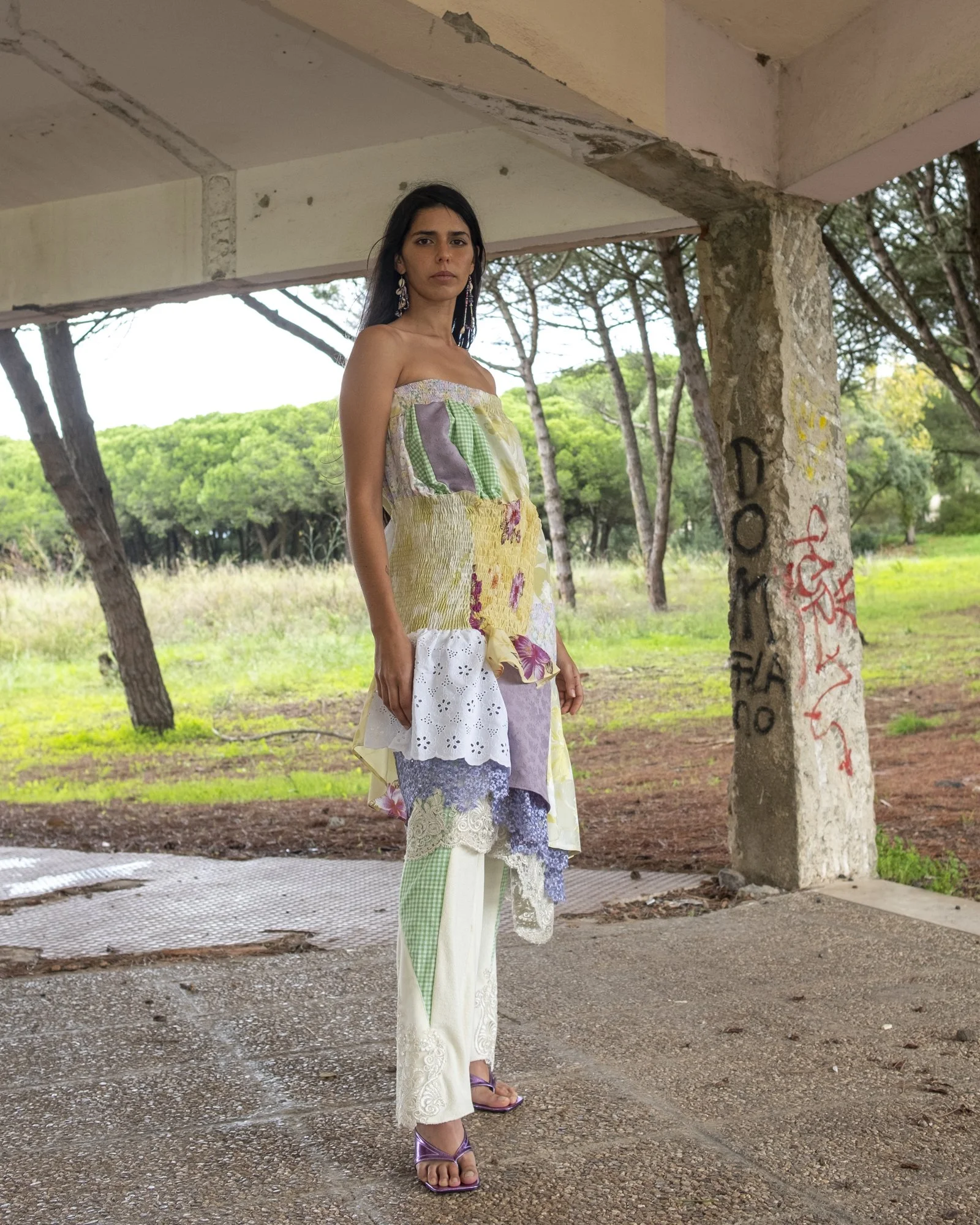 A woman wearing a colorful patchwork dress with lace and eyelet details stands outdoors under an overpass with graffiti on a concrete pillar, with a green wooded area in the background.