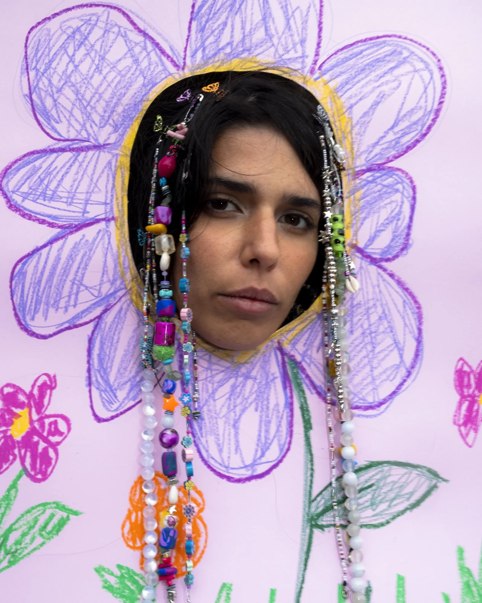 Person with dark hair and neutral expression looking through a flower-shaped cutout in a pink drawing of a flower with purple petals and green leaves on a pink background decorated with added colorful bead necklaces.
