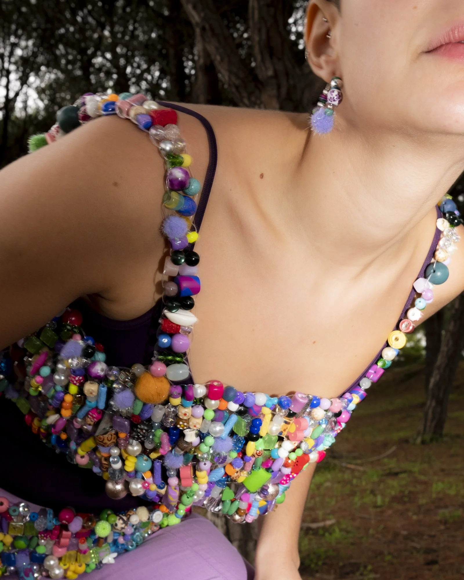 Close-up of a woman wearing a colorful beaded top and earrings, outdoors with trees in the background.