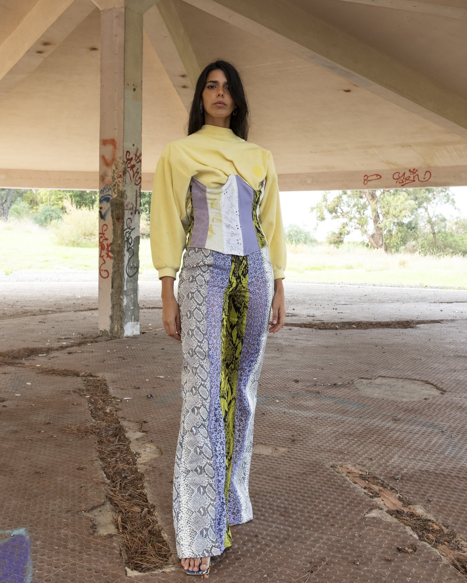 A woman standing under a concrete structure with graffiti, wearing a yellow sweatshirt with a corset-style top and wide leg printed pants with snakeskin and animal print patterns.