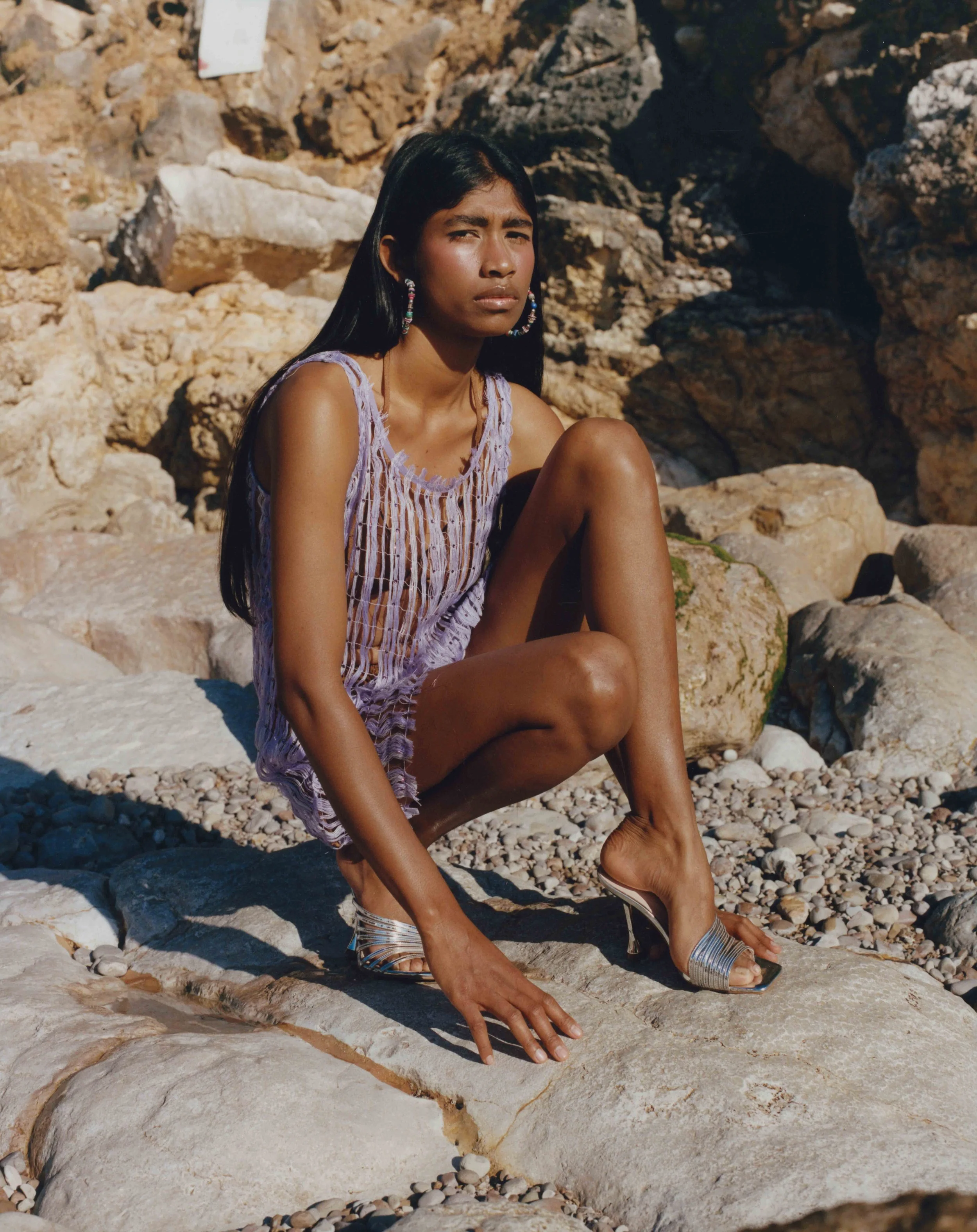 A woman sitting on rocks at a beach, wearing a light purple dress and high heel sandals, with a serious expression, sunlight, and rocky background.