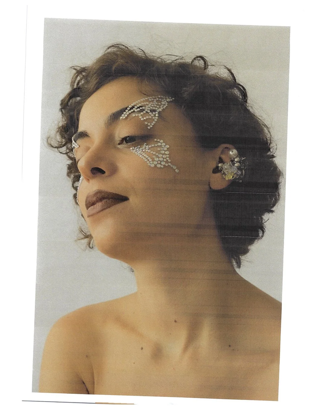 A woman with short, curly hair and bold makeup, wearing rhinestone face art around her eyes and large earrings.