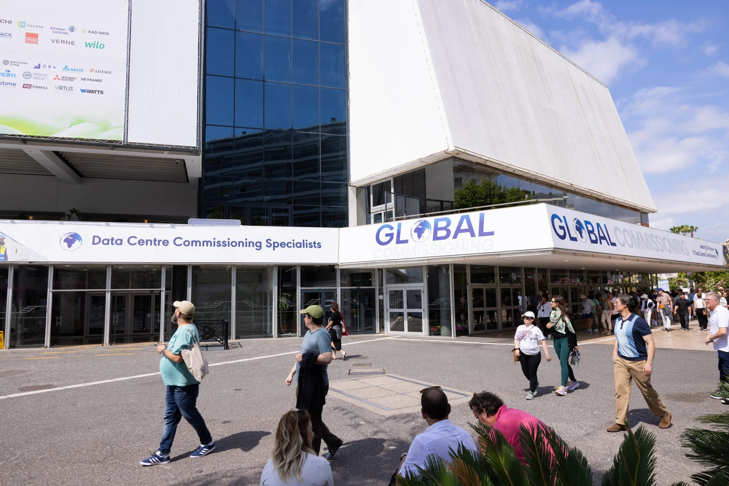 People walking outside the Data Centre Commissioning Specialists event at a modern building with signage that reads 'Global', 'Data Centre Commissioning Specialists', and 'Global Commissioning' on the front.