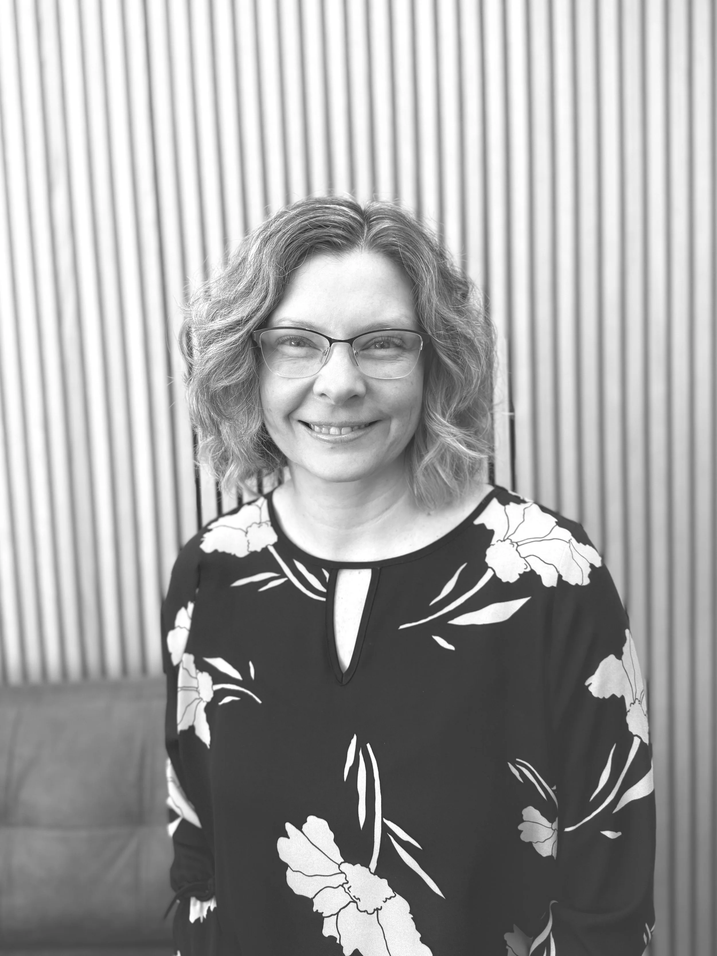 A woman with curly hair and glasses smiling, wearing a floral-patterned top, standing in front of a vertically striped wall.