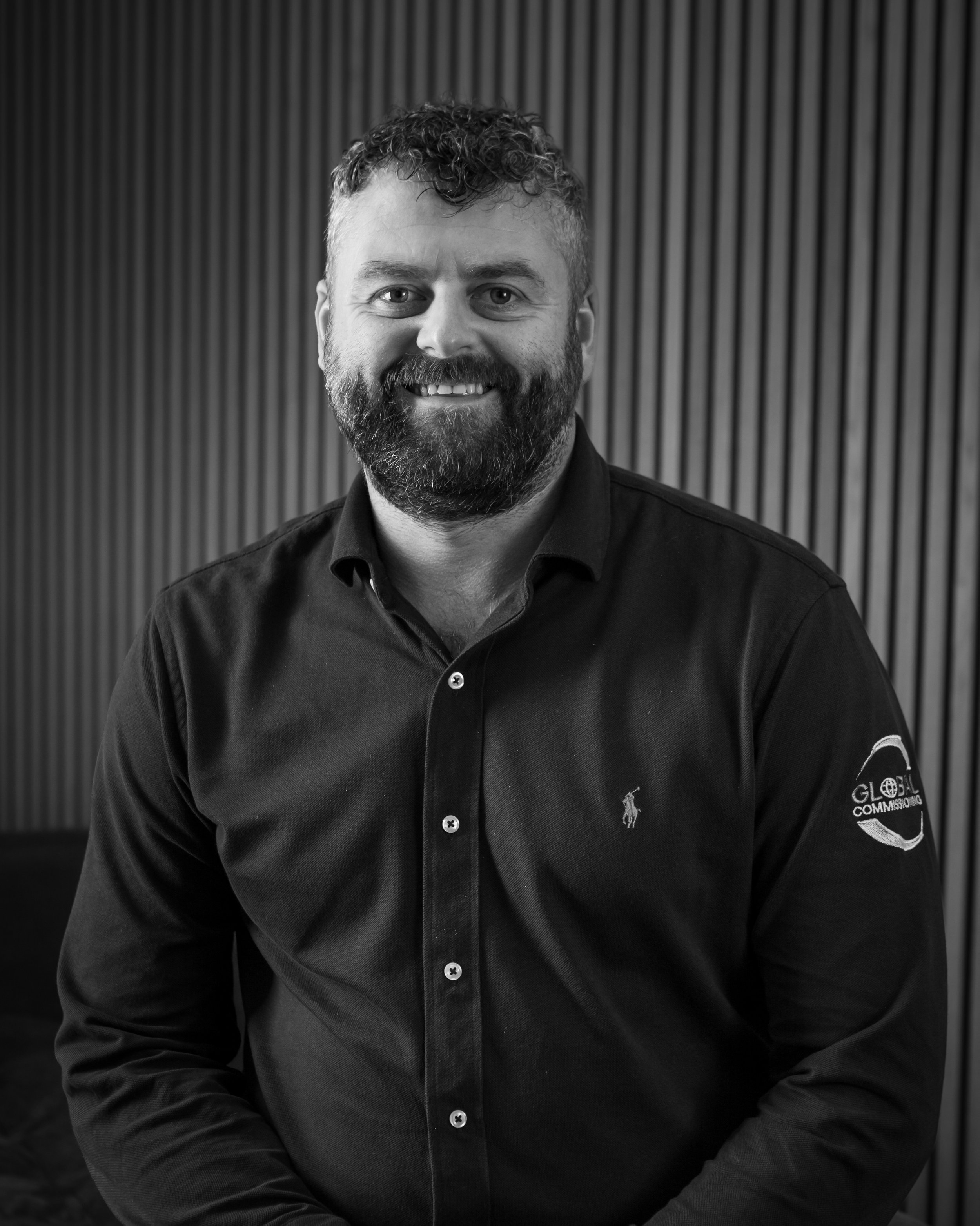A man with curly hair and a beard smiling, wearing a black shirt with a logo on the sleeve, standing in front of a textured wall.