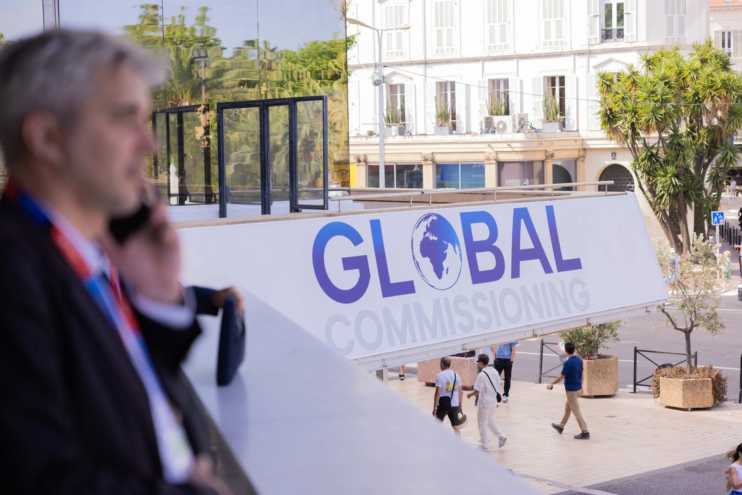 A blurred man speaking on a phone in the foreground, with a large sign that reads 'Global Commissioning' and a globe logo in the background. Several people are walking outside on a sunny day, near a building with white walls, balconies, and potted plants.