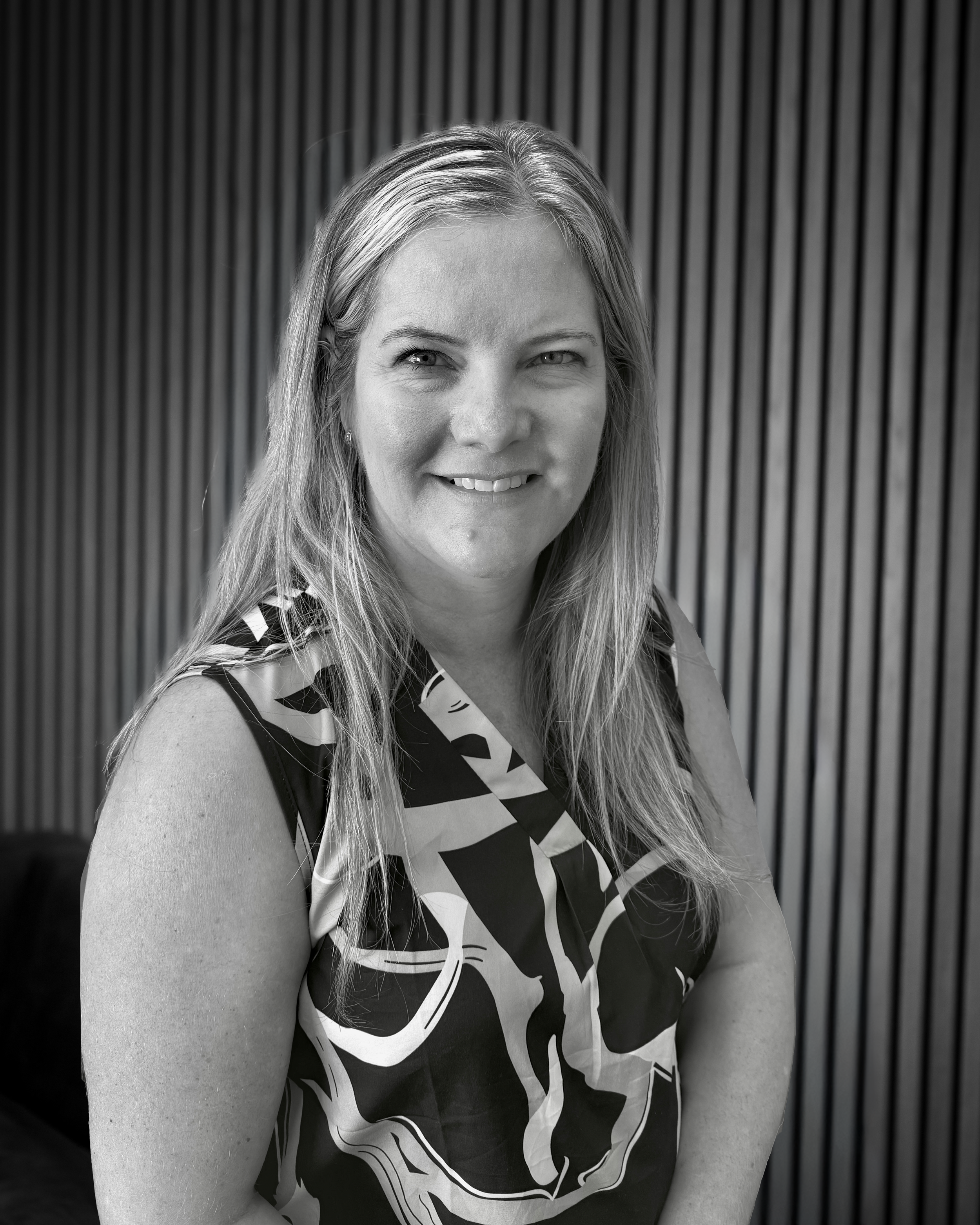 A woman with long hair smiling at the camera, wearing a sleeveless top with a bold, abstract pattern, sitting in front of a vertical wooden slat wall.