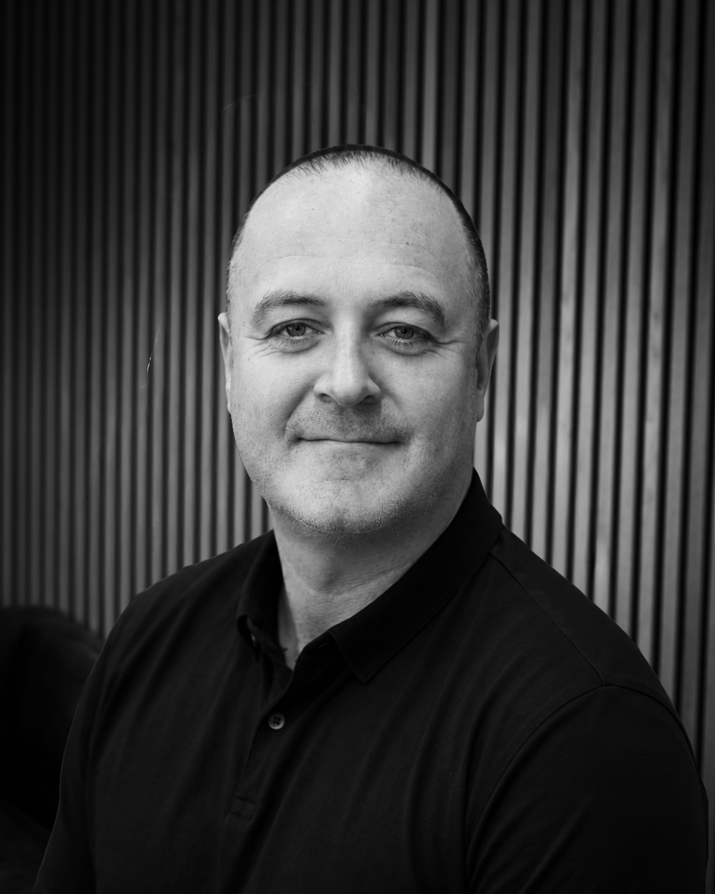Black and white portrait of a smiling man in a black button-up shirt with a polo logo on the chest, standing against a background with vertical wooden paneling.