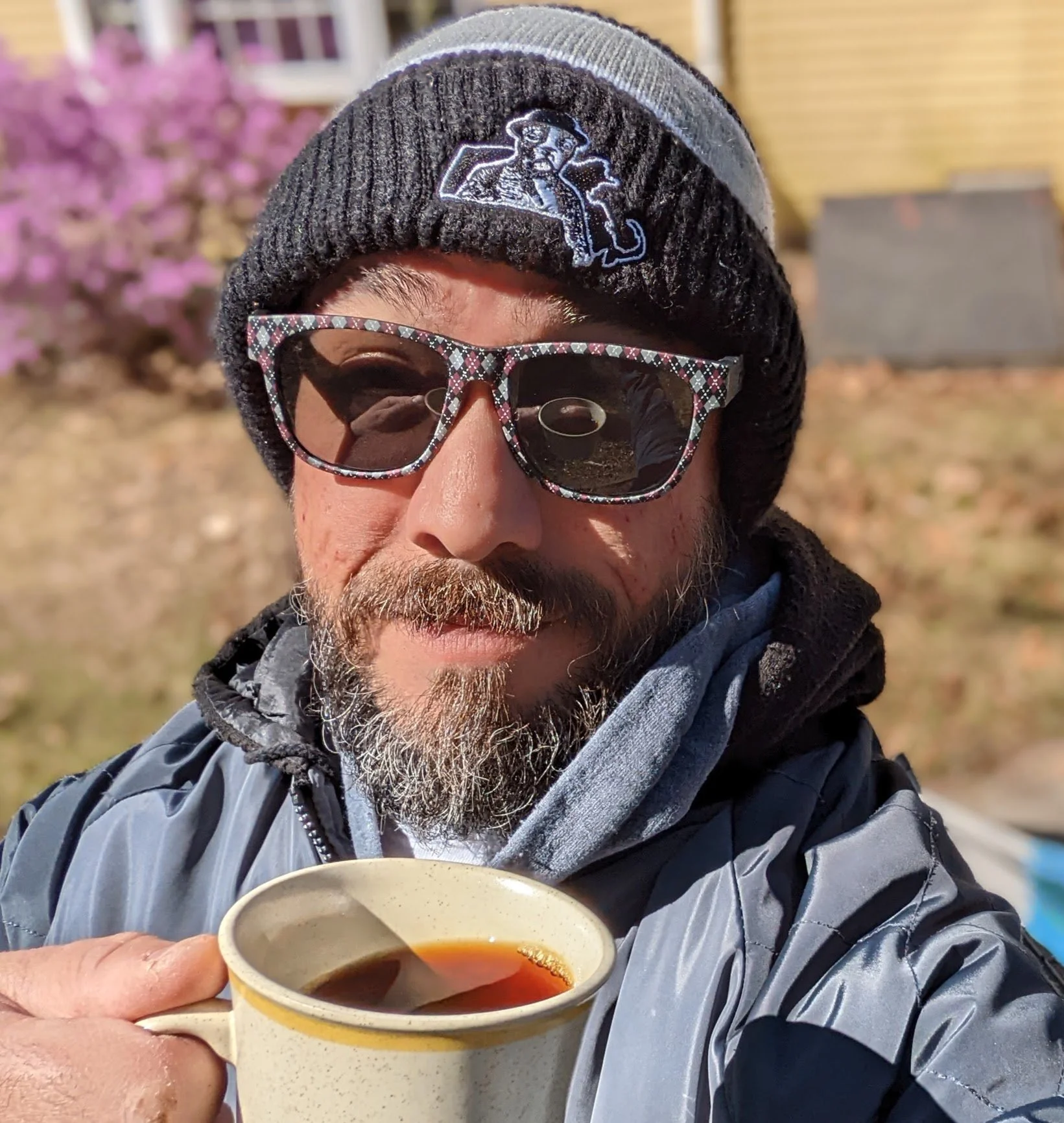 A person wearing a black beanie with a logo, sunglasses with a patterned frame, and outdoor clothing, holding a cup of tea or coffee outdoors with pink flowering shrub and building in the background.