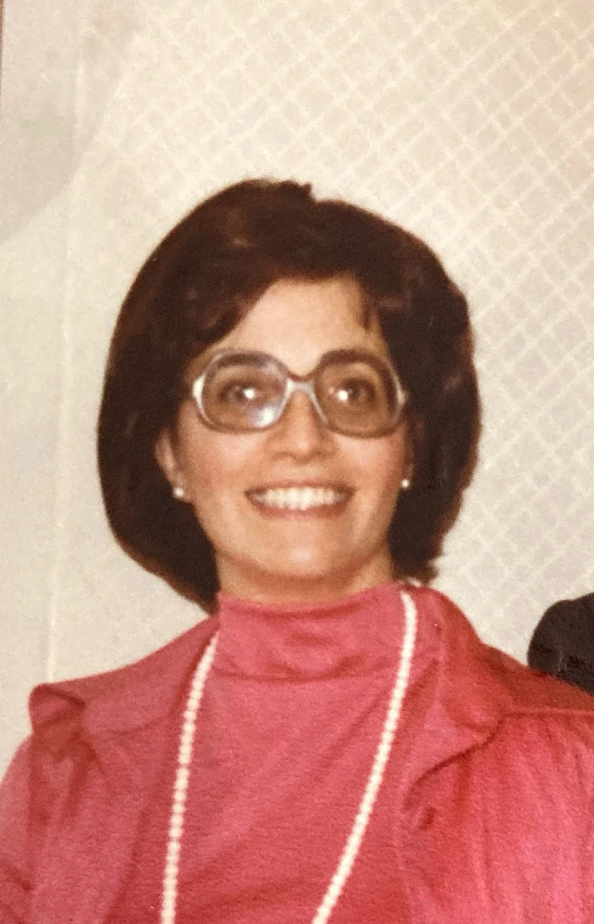 A woman with dark hair, wearing glasses, earrings, a pink turtleneck, and a pink blazer with a white trim, smiling at the camera.
